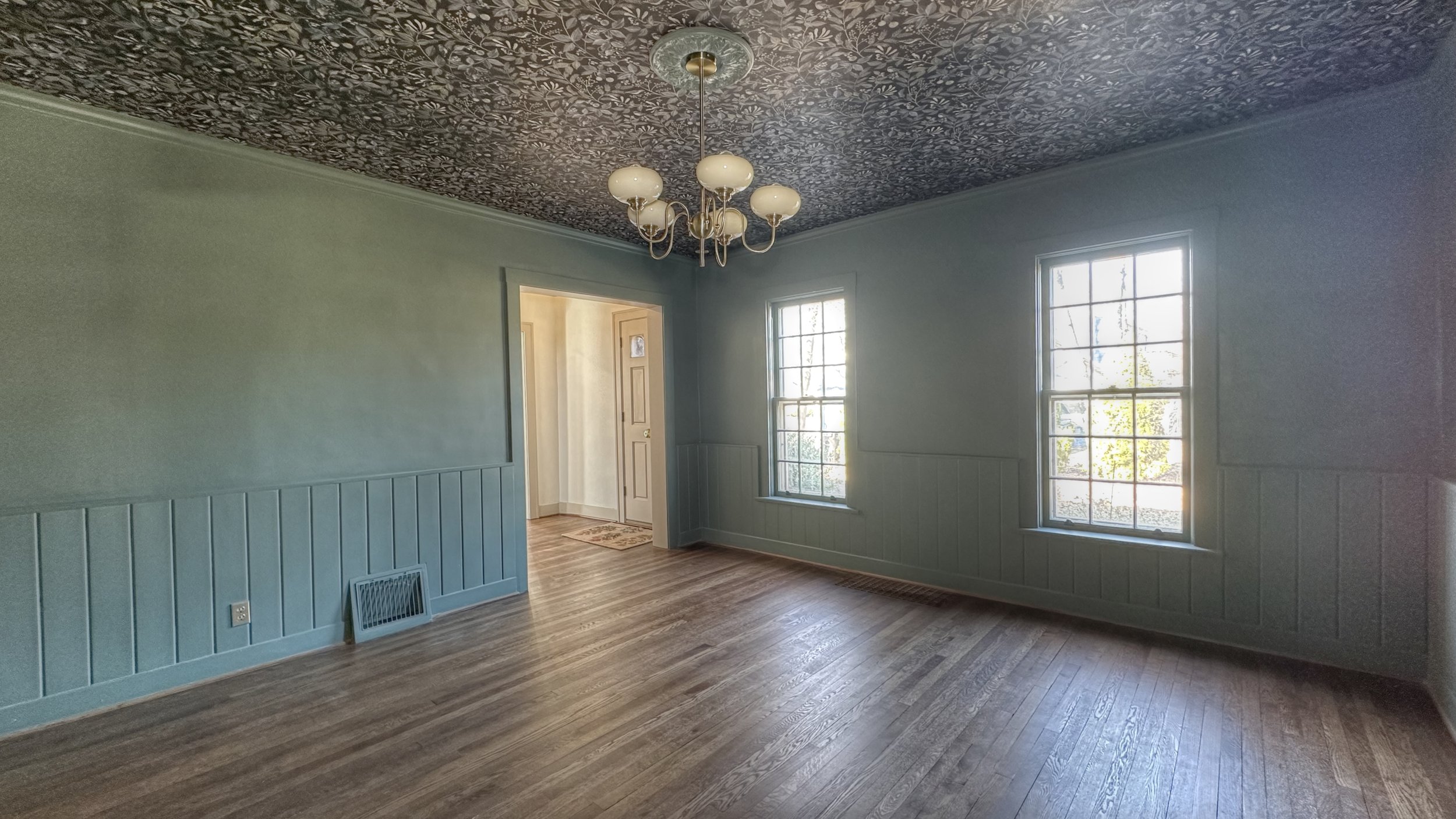 Dining room with hardwood floors, two large windows, and soft blue walls with decorative lower wall paneling. A vintage-style chandelier hangs from a patterned ceiling, and an open doorway leads to an adjacent room.
