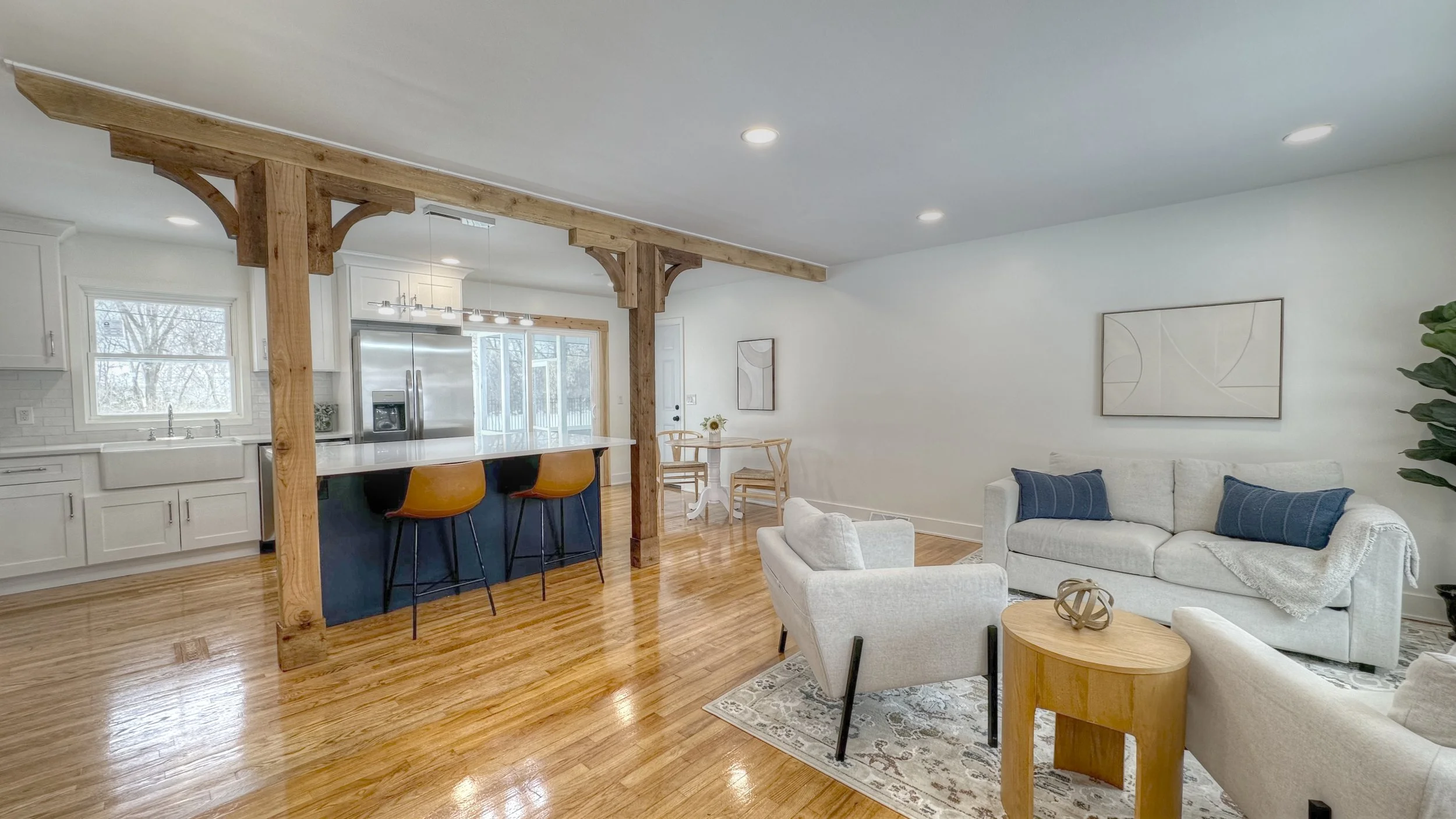 Open-concept living room and kitchen with hardwood floors, white walls, a sofa with blue pillows, a white armchair, a round wooden side table, a small dining table with chairs, and a kitchen with white cabinets and a large stainless steel refrigerator. Wooden beams add architectural detail.
