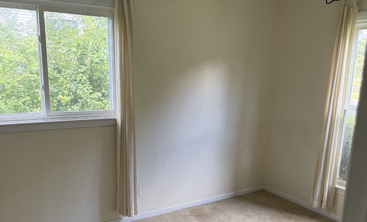 Empty bedroom with beige carpet, light walls, and two windows with curtains before renovation