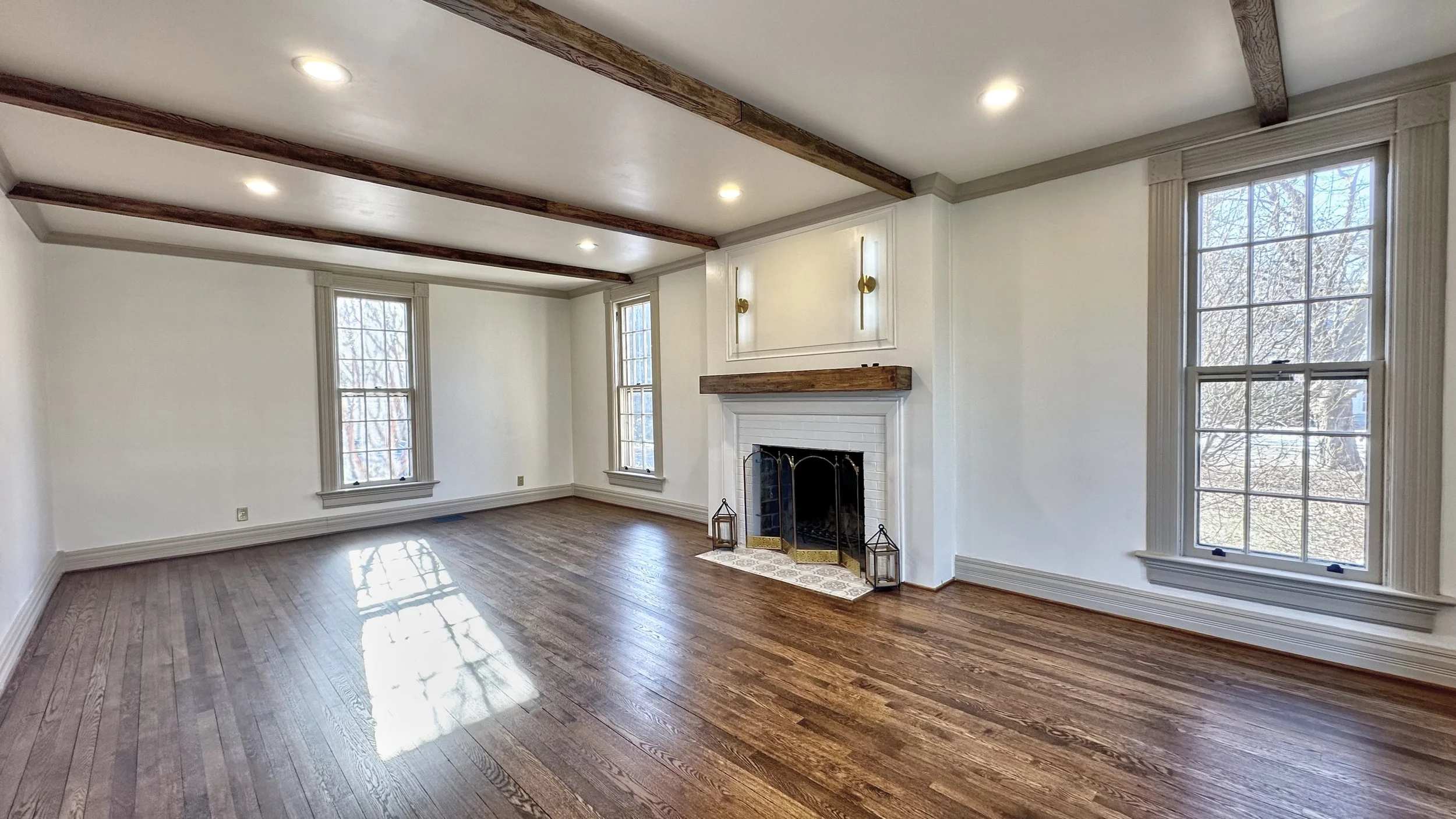 Large, sunlit living room with rich hardwood floors, exposed wood ceiling beams, and a white brick fireplace with a wood mantel. Multiple tall windows bring in natural light, highlighting the open layout and classic architectural details