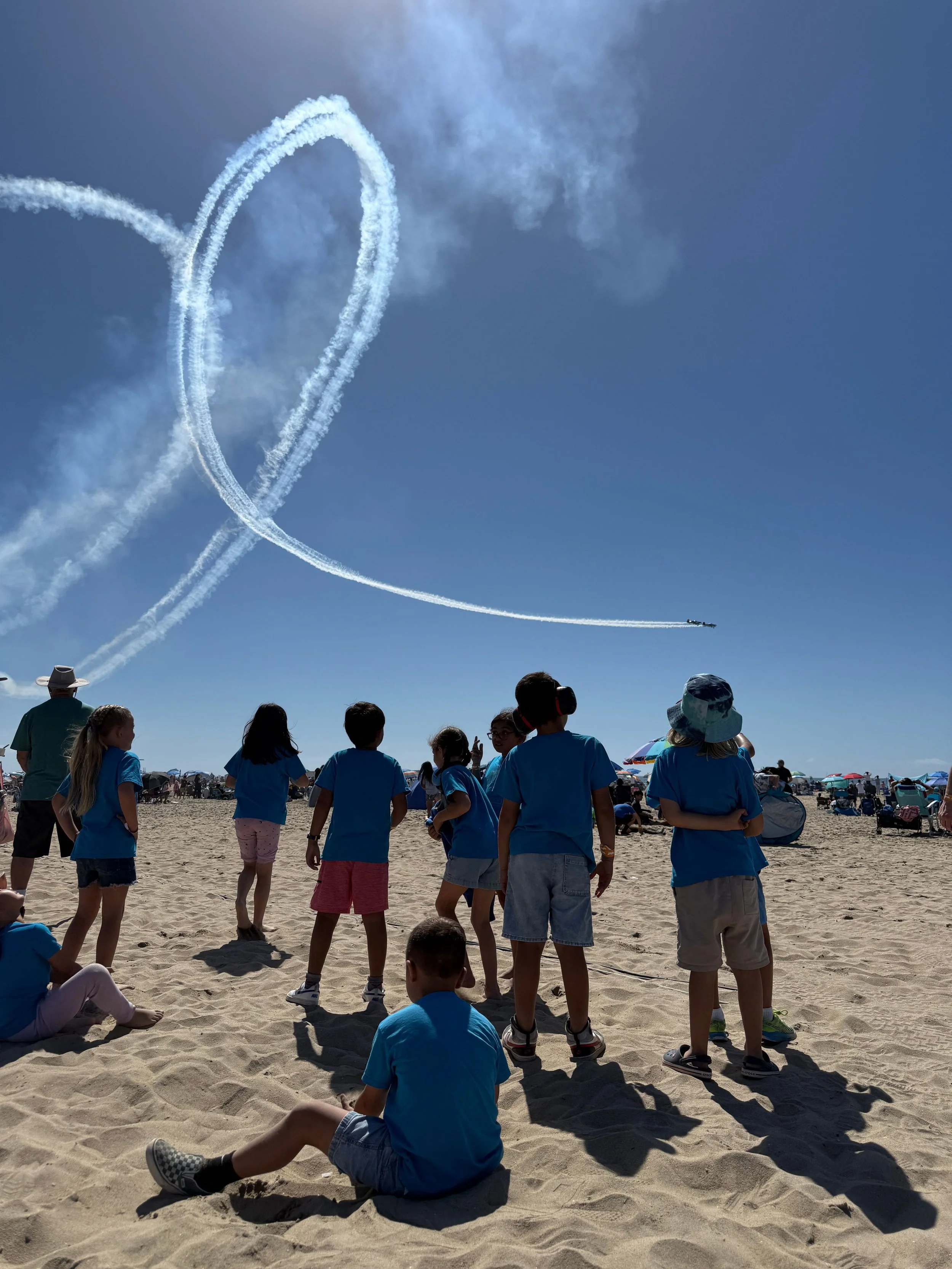 Children and adults on a sandy beach watch a plane perform an air show, creating heart-shaped smoke trails in the blue sky.