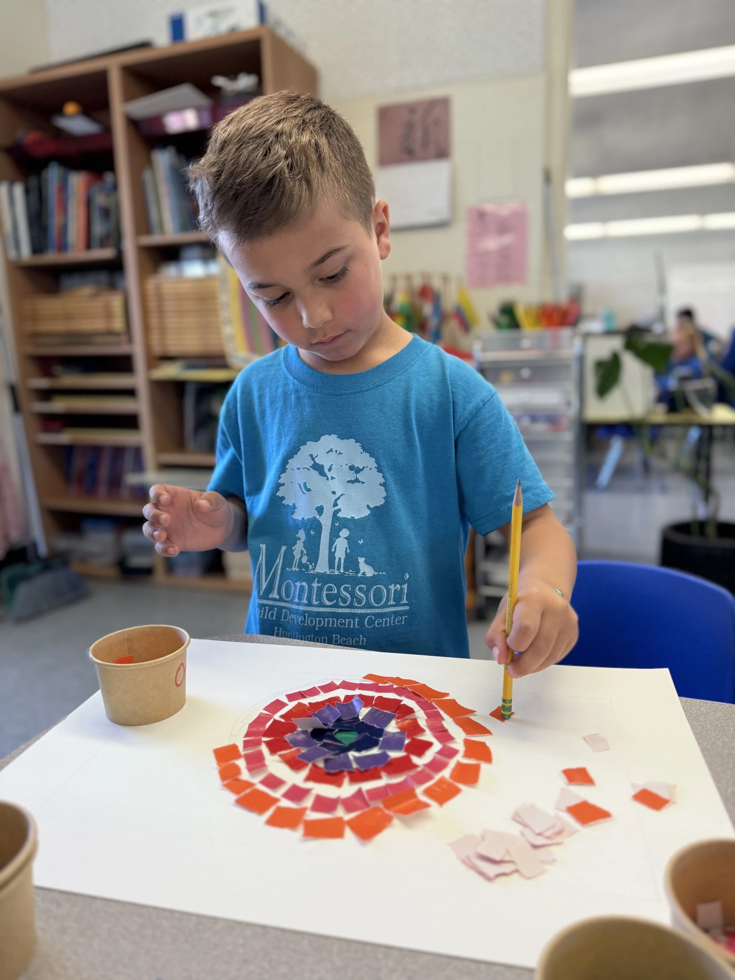 A young boy in a blue Montessori t-shirt working on a colorful mosaic art project in a classroom, with shelves filled with books and educational materials in the background.