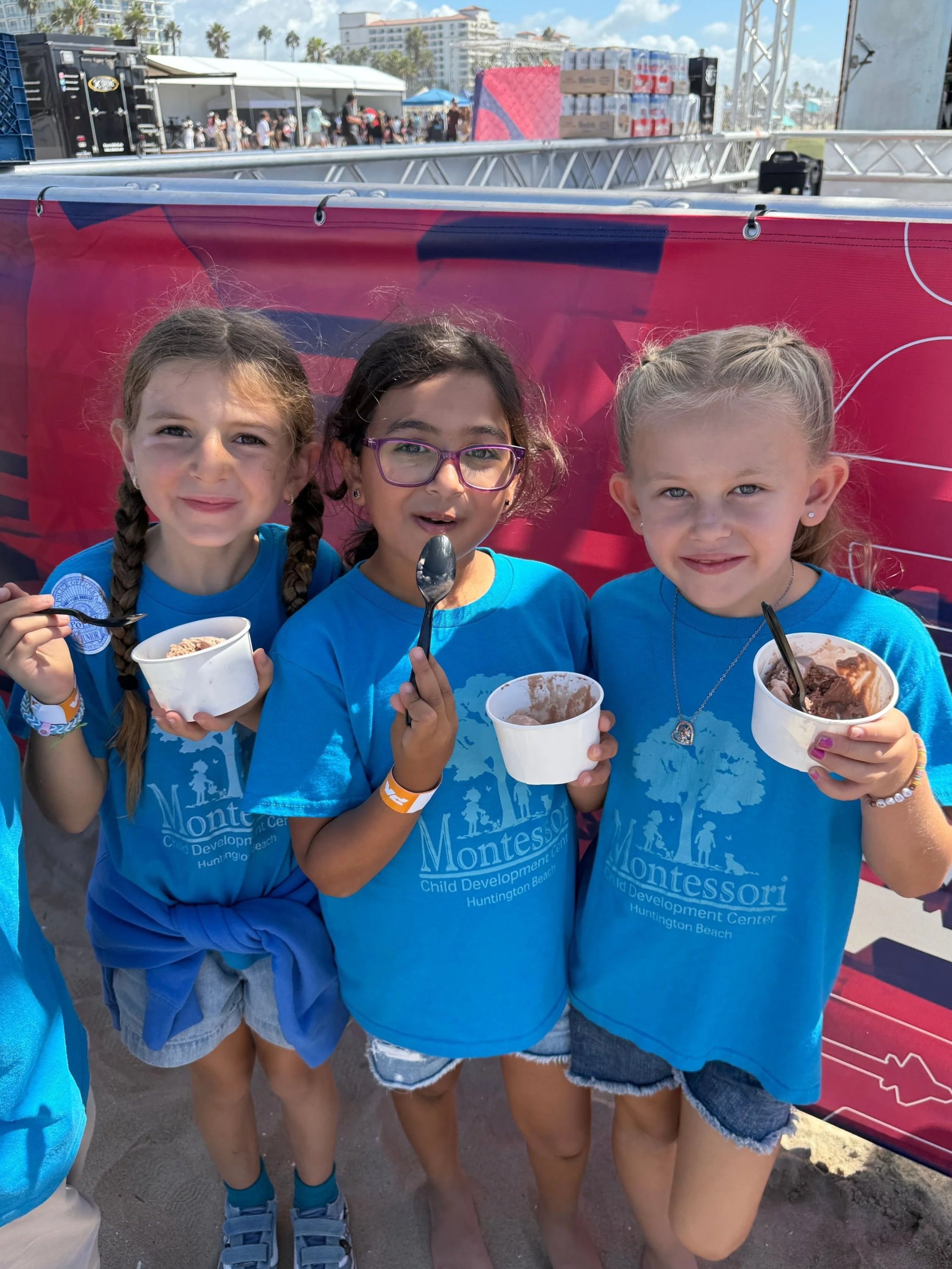 Three young girls in blue Montessori T-shirts standing on the beach, holding bowls of ice cream and smiling at the camera.