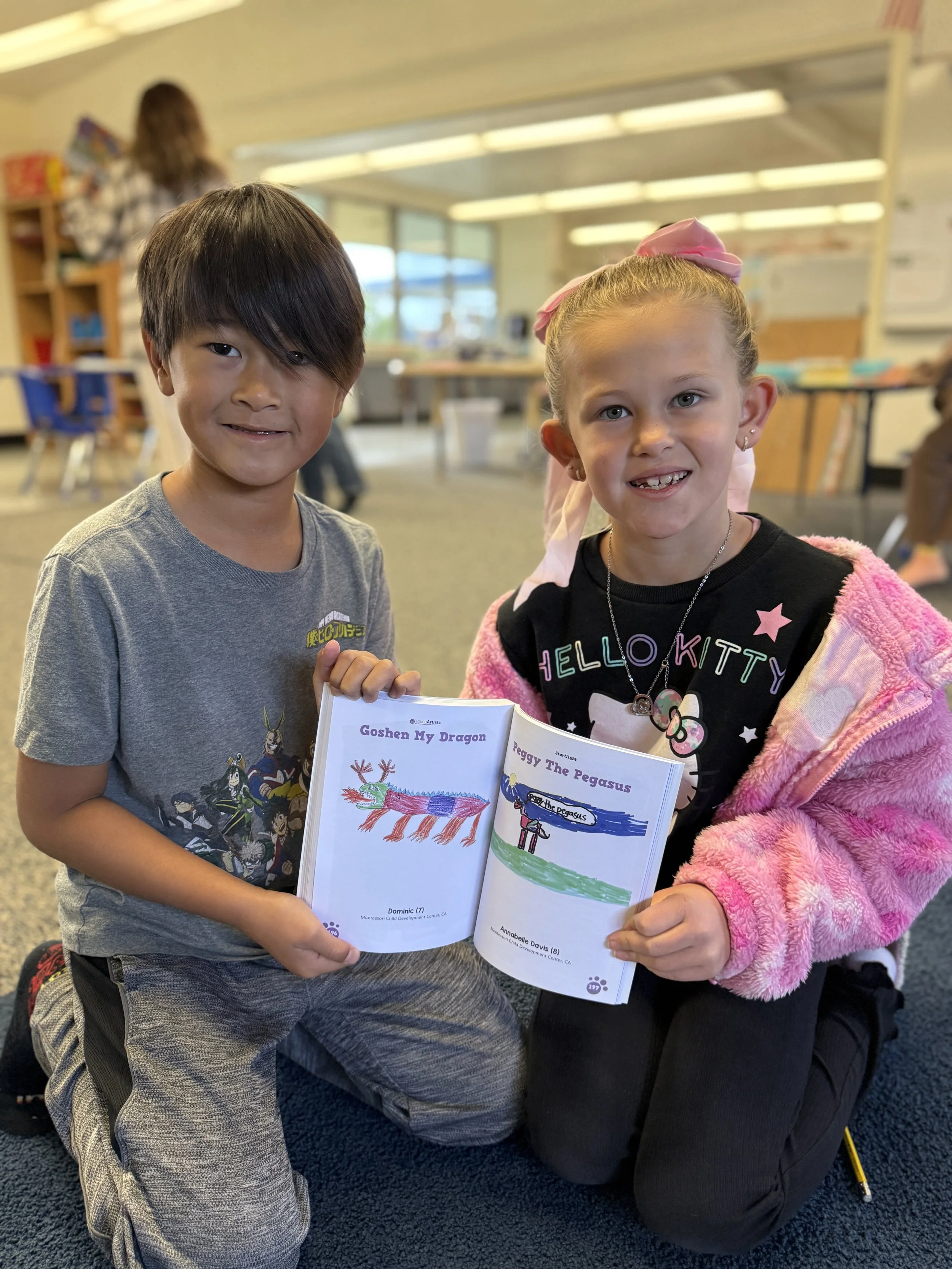 Two children kneeling on a carpeted floor in a classroom, holding up an open book with colorful drawings and text. The boy on the left has black hair and is wearing a gray T-shirt, while the girl on the right has blonde hair with a pink bow and is wearing a black 'Hello Kitty' shirt with a pink jacket.