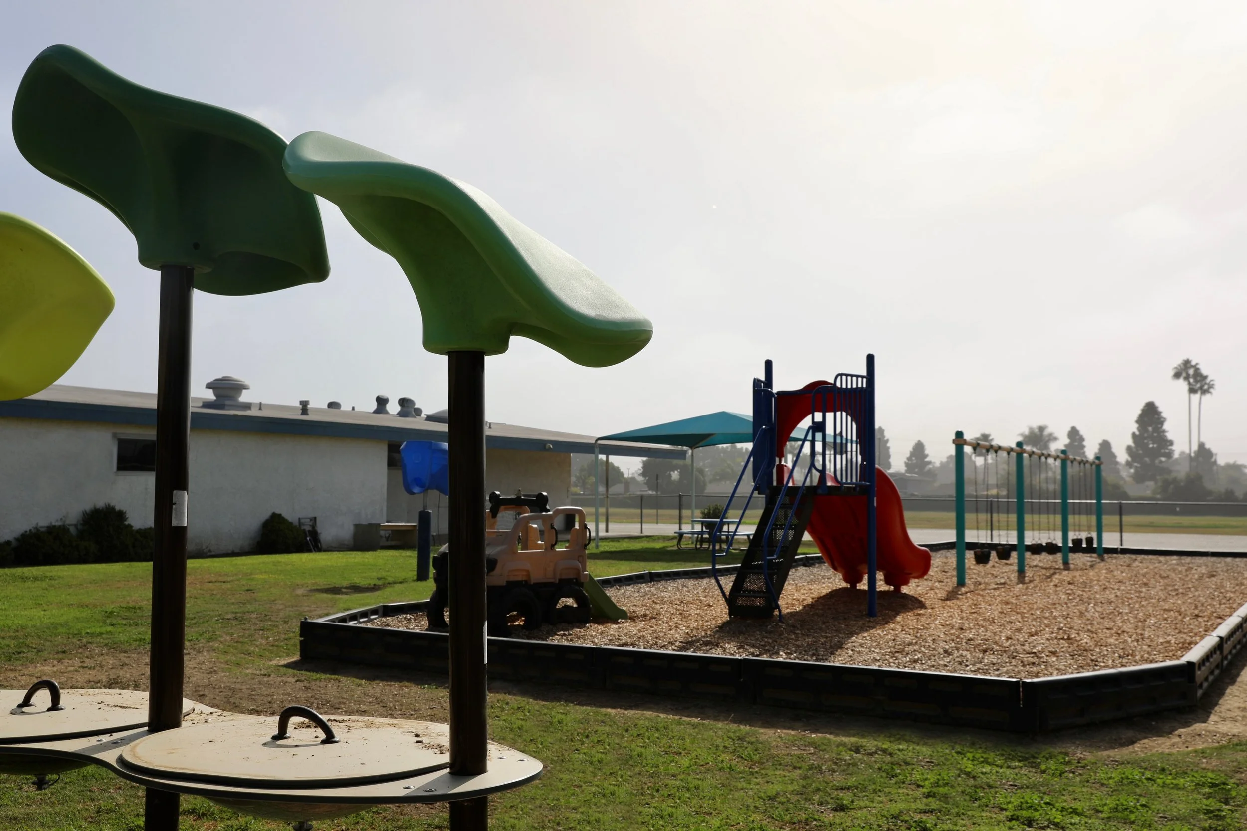 Children's playground with green shade structures, a red slide, a see-saw, and swings on a gravel surface, with a building and trees in the background under a cloudy sky.