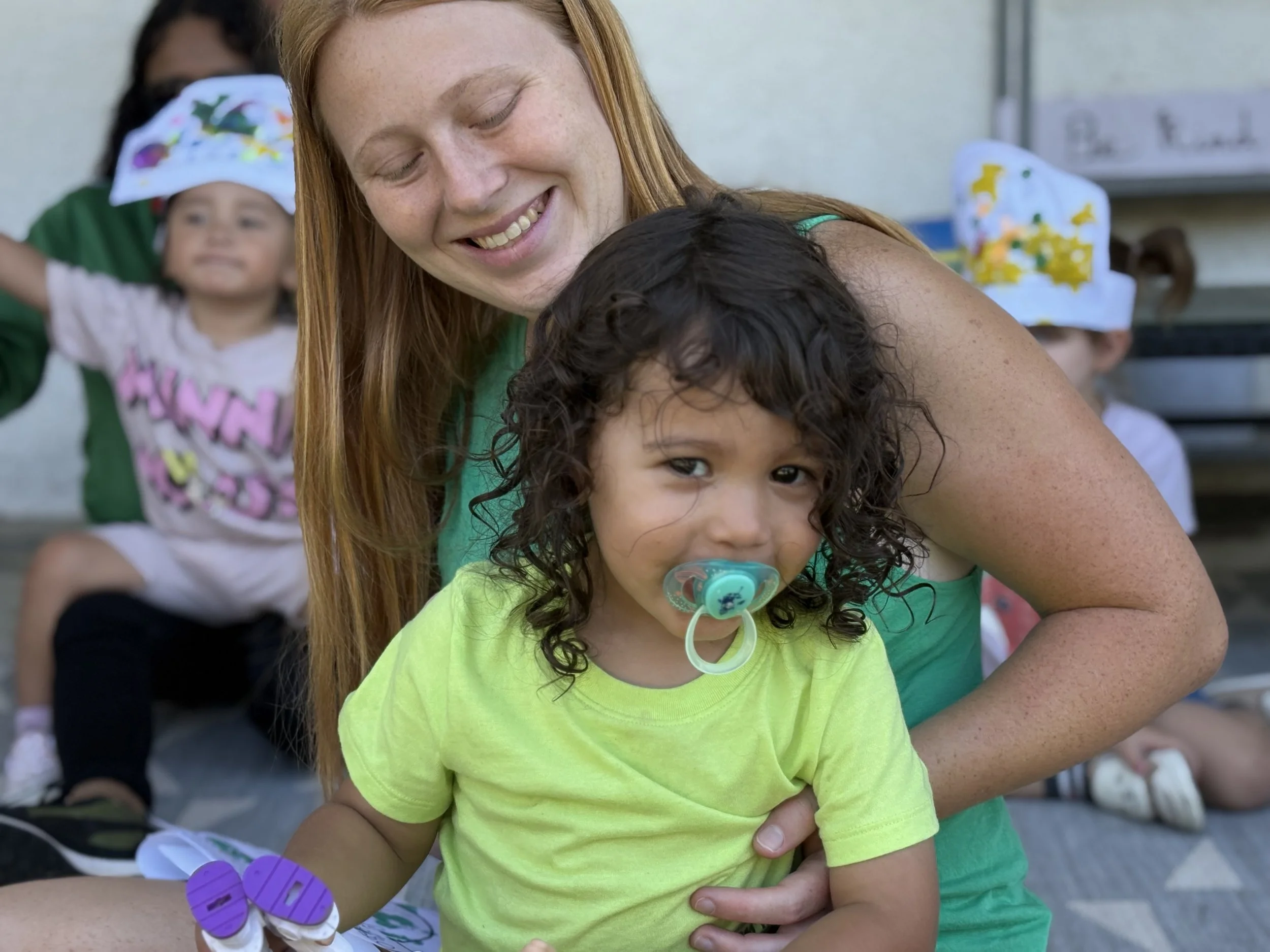 A woman with red hair holding a young girl with curly hair and a pacifier, with other children in the background at an outdoor event.