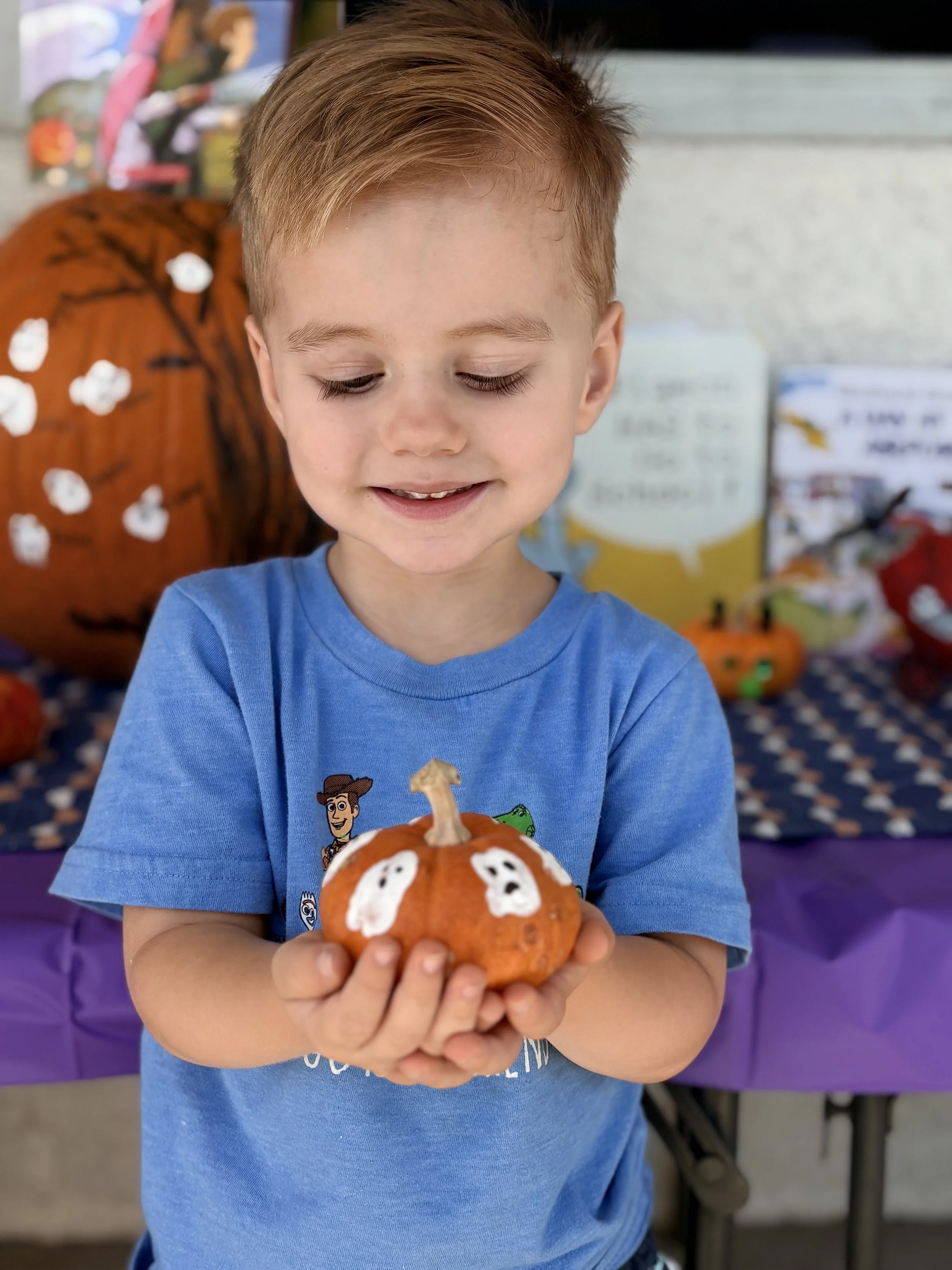A young boy holding a small decorated orange pumpkin with white ghost shapes, at a Halloween-themed event.