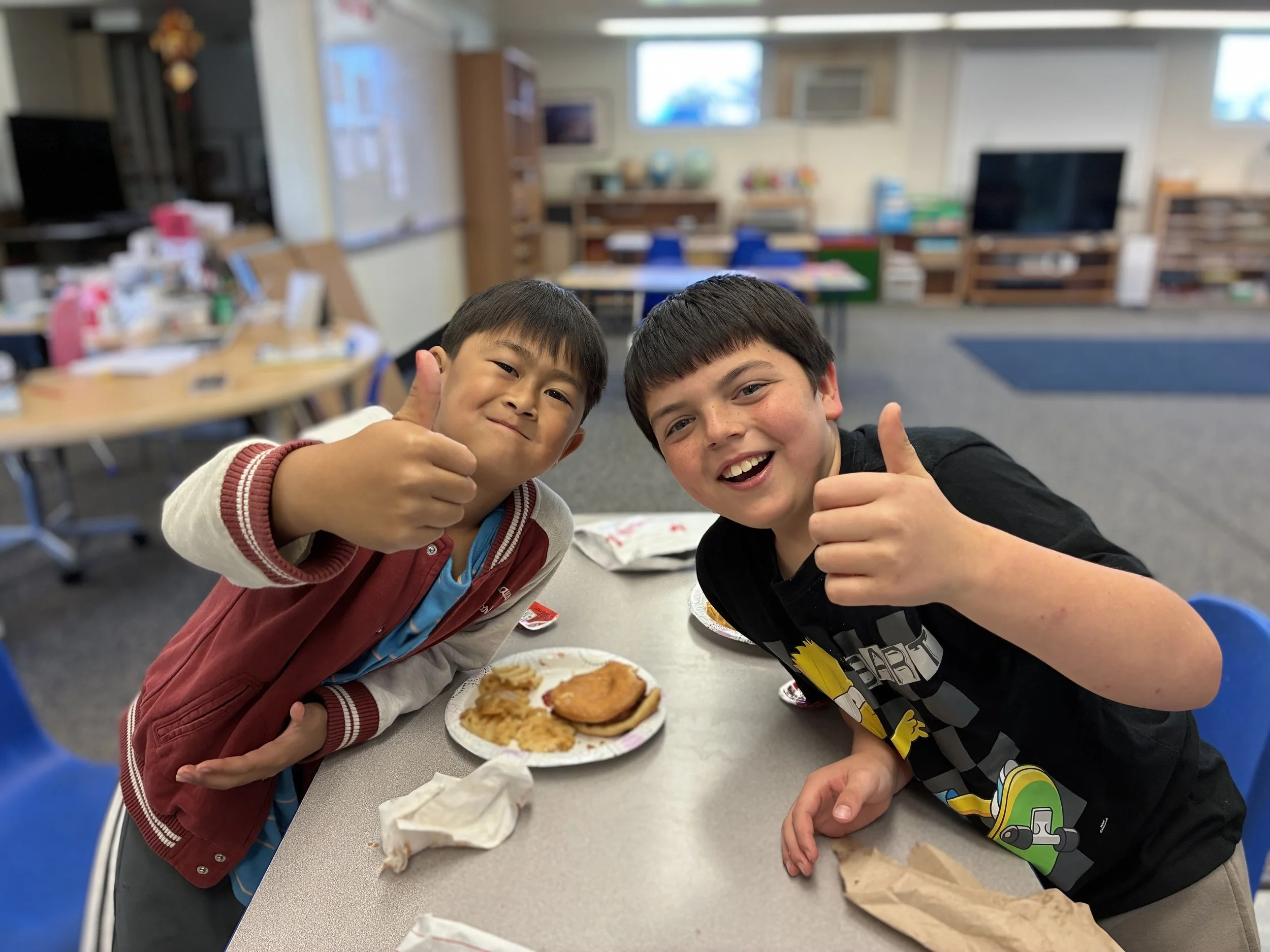 Two boys smiling and giving thumbs up at a table with food in a classroom.