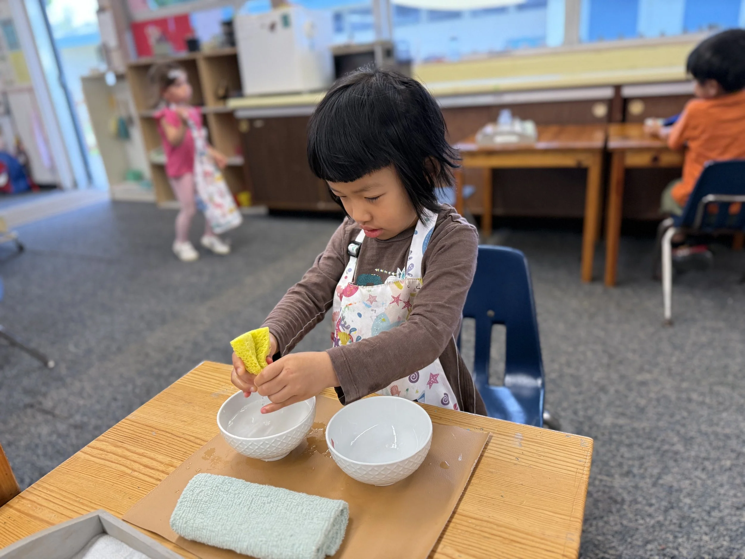 A young boy wearing an apron washing a yellow sponge in one of two white bowls on a wooden table in a classroom setting.
