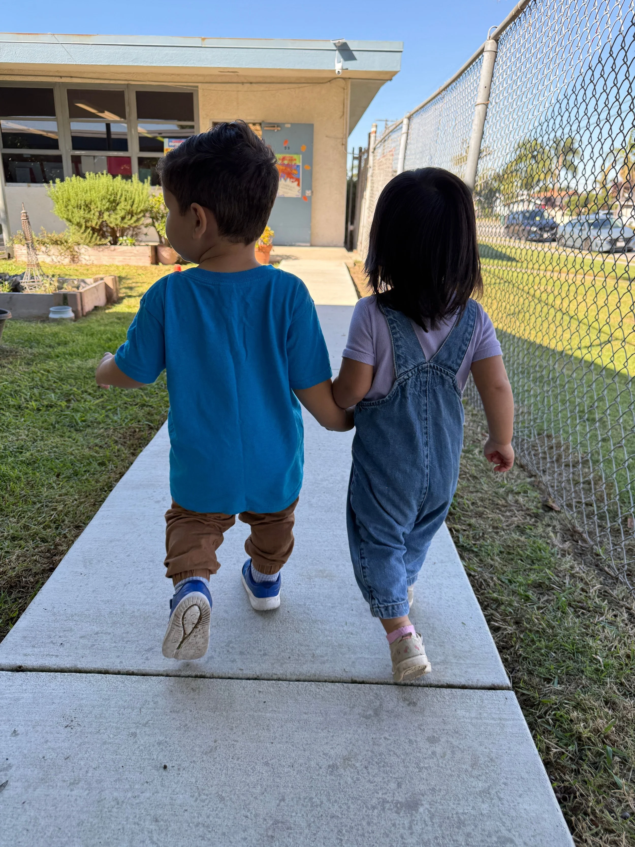 Two young children, a boy and a girl, walking hand in hand on a sidewalk outside a building with a chain-link fence on the right and a grassy area on the left.