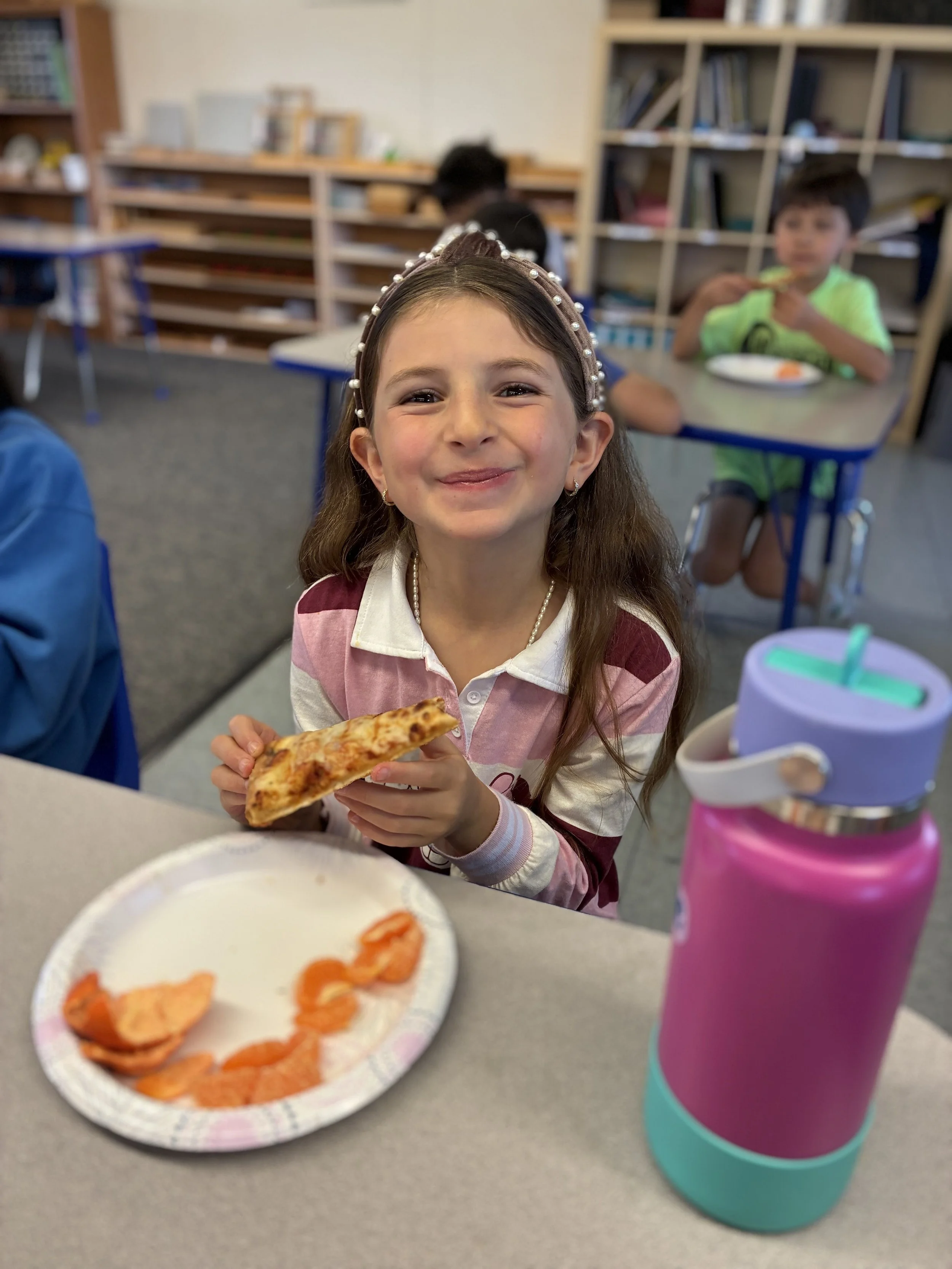 A young girl with long brown hair wearing a pearl headband and a pink and white striped shirt is smiling and holding a slice of pizza in a classroom. There is a paper plate with orange slices and a pink water bottle on the table in front of her. Other children and bookshelves are visible in the background.