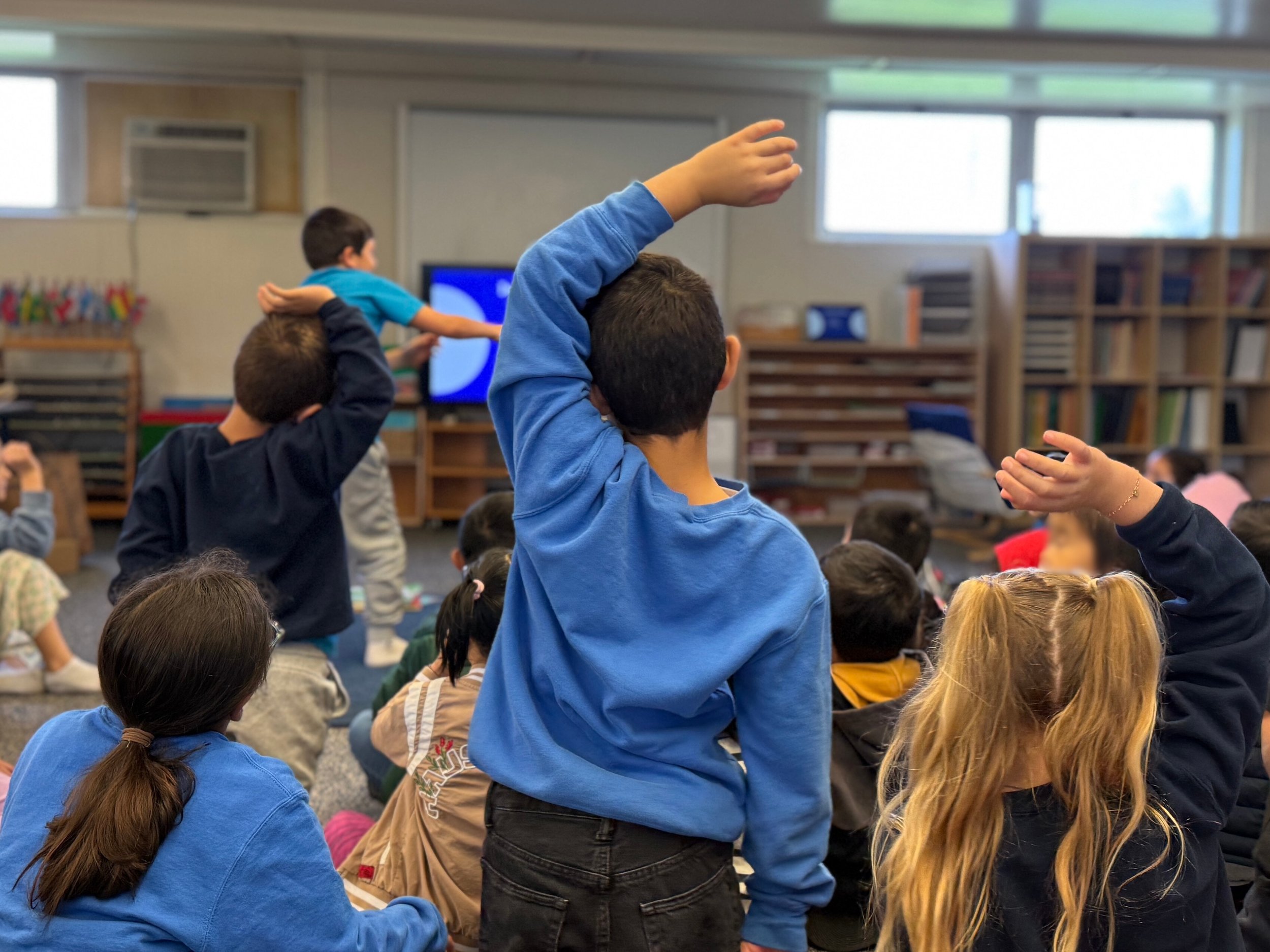 Children in a classroom, some raising their hands, seated on the floor, with a teacher or presenter at the front, and bookshelves in the background.