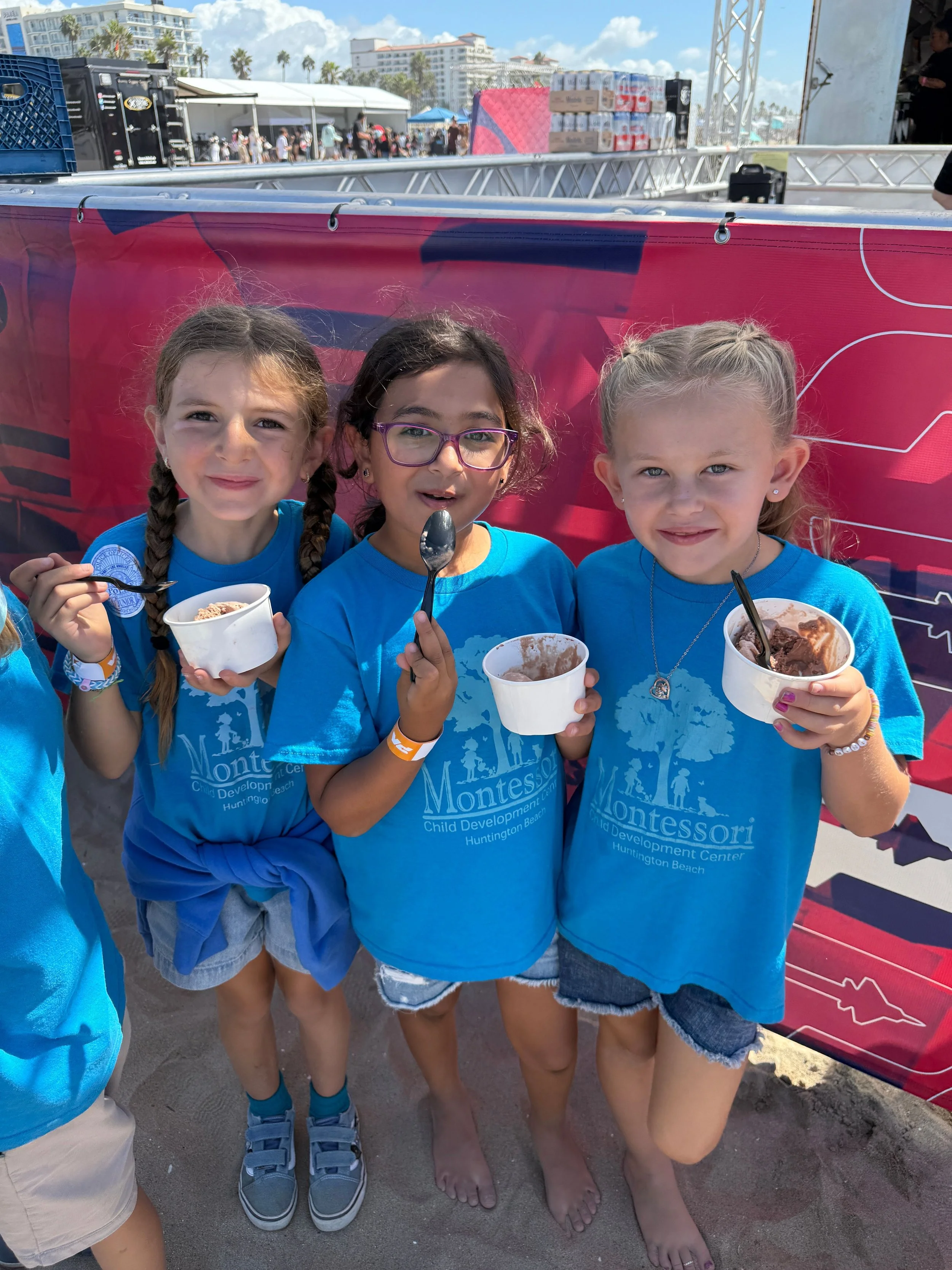 Three young girls in blue Montessori T-shirts standing on the beach, holding bowls of ice cream and smiling at the camera.