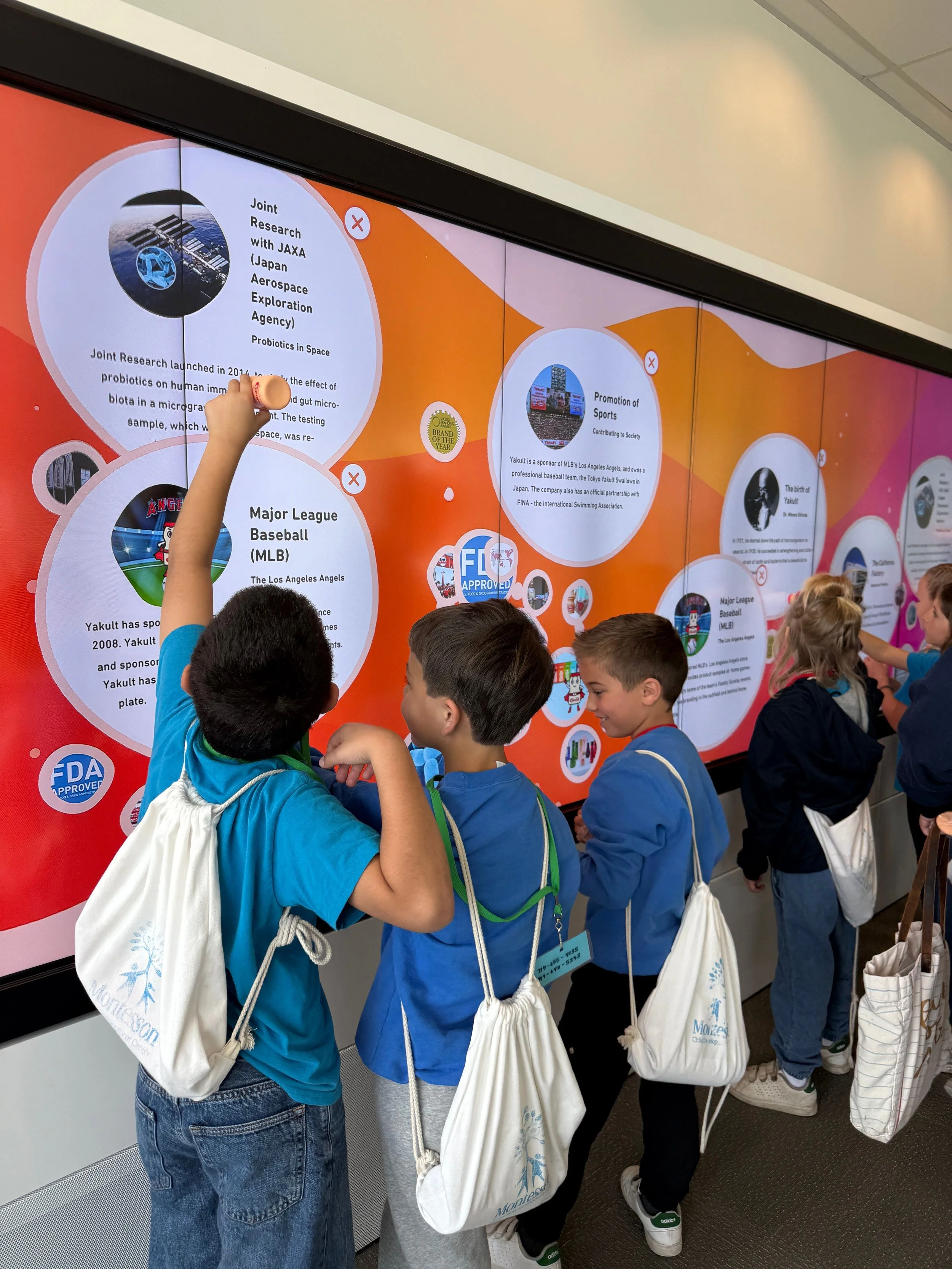 Children in blue shirts with white drawstring bags look at a large digital display screen with colorful informational circles, including NASA and sports content, at an educational exhibit or museum.