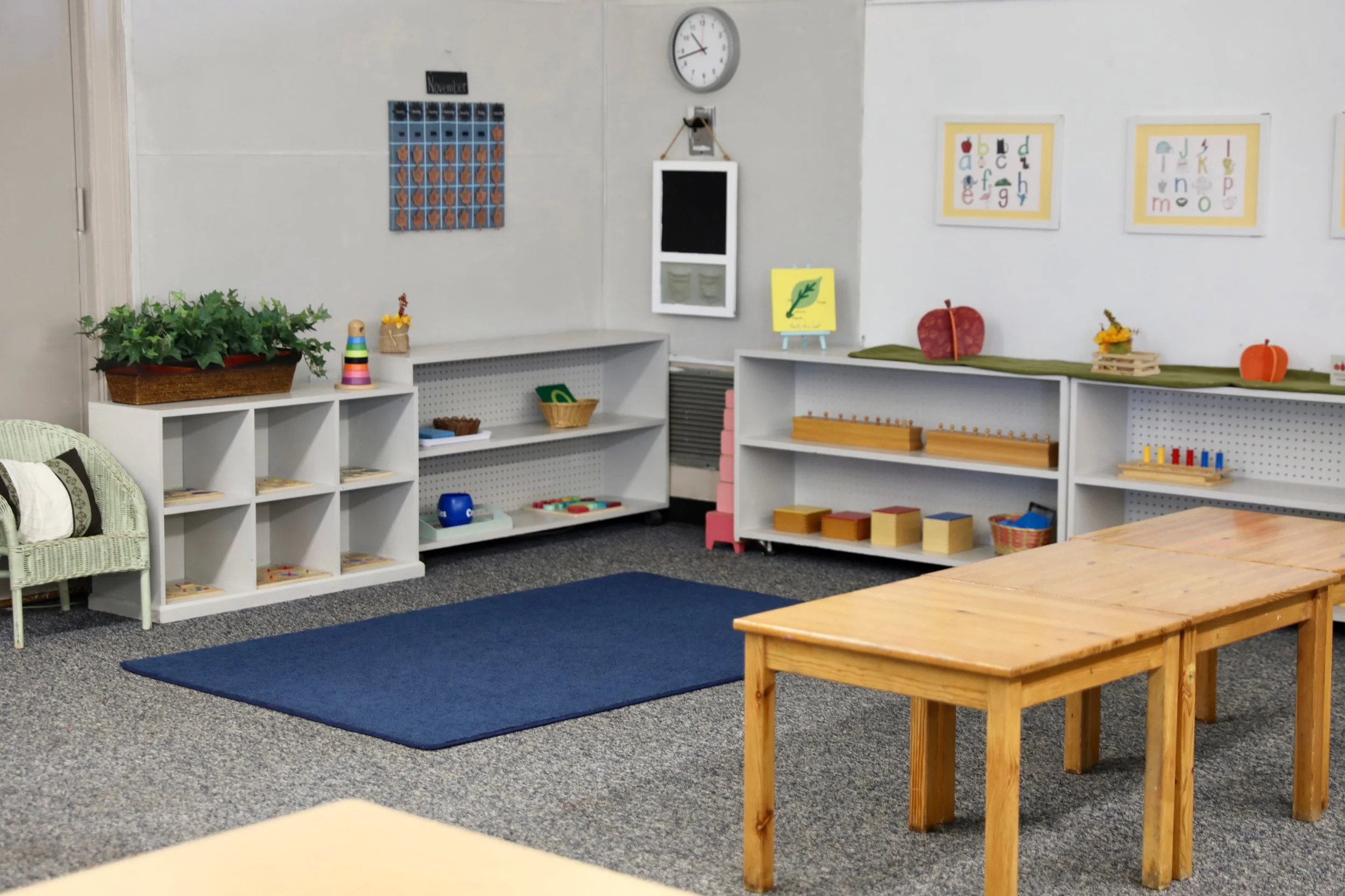 A classroom with bookshelves, puzzles, and educational decorations, including a clock and alphabet pictures on the walls, along with a blue rug and wooden tables.
