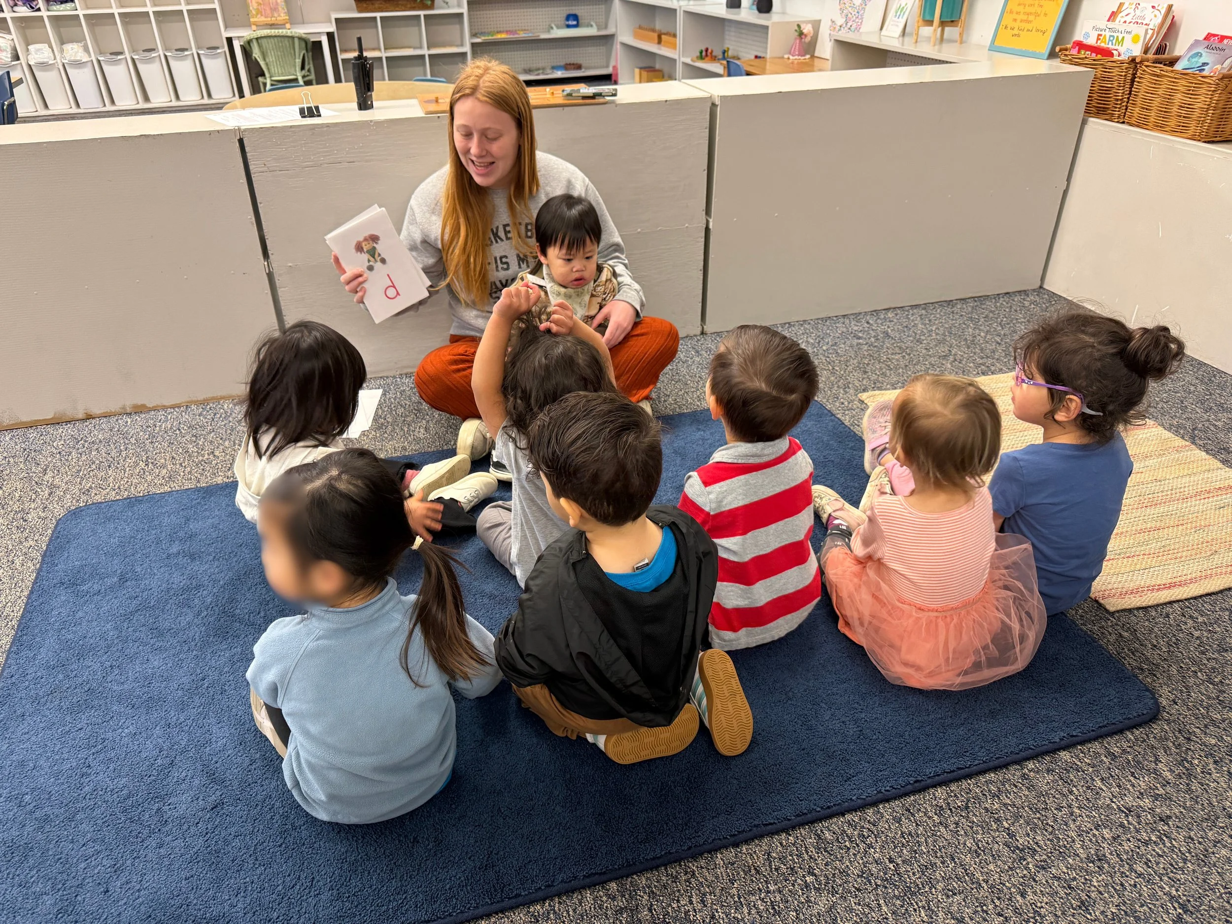 A group of young children sitting on a blue carpet in a classroom, gathered around a teacher holding a book with a letter 'p' on the cover. The teacher appears to be engaging the children in a story or lesson.