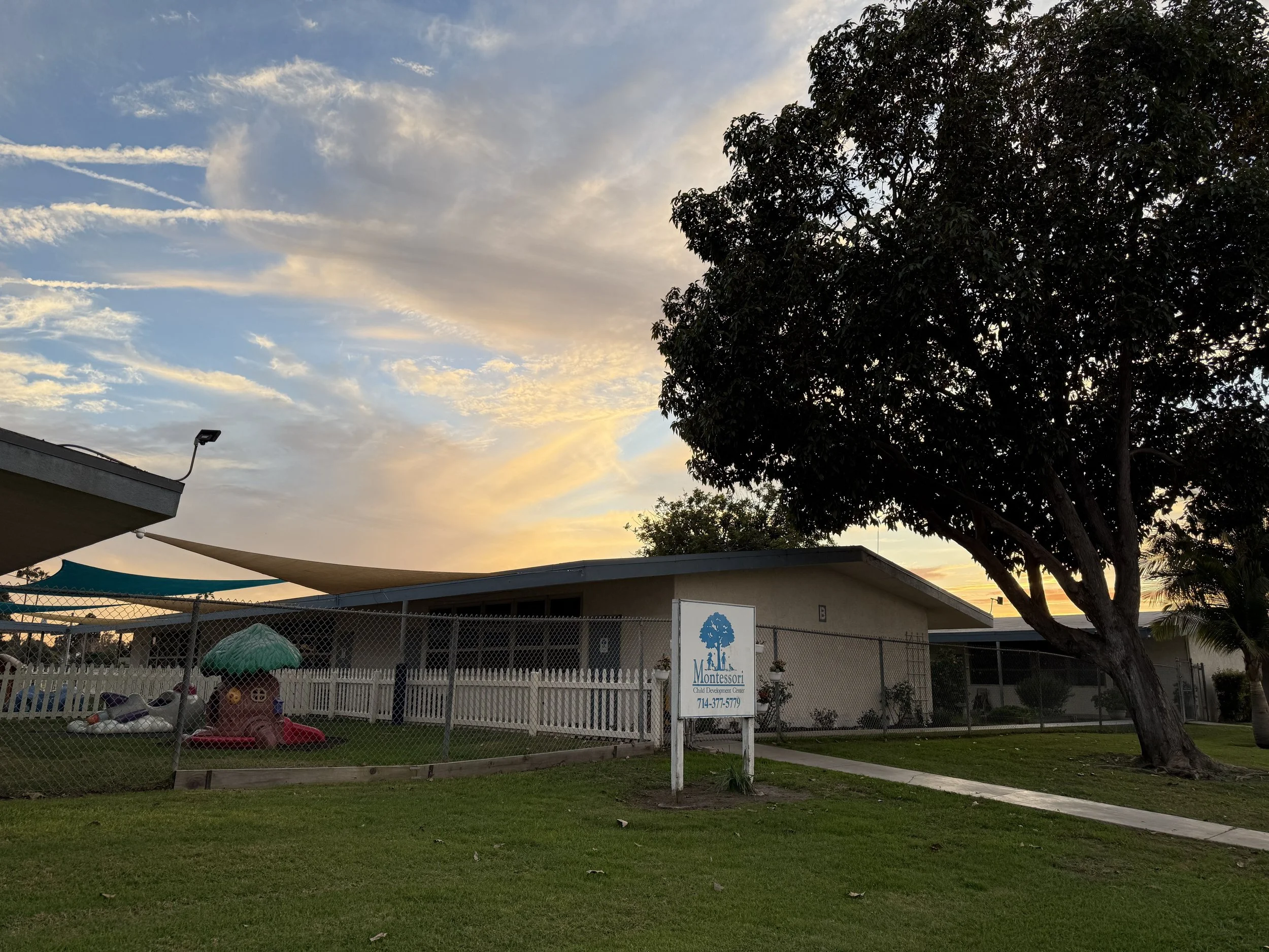 A preschool playground enclosed by a chain-link fence with a sign reading 'Montessori Chil Preschool' and a phone number, with a large tree, a grassy yard, and a sunset sky in the background.