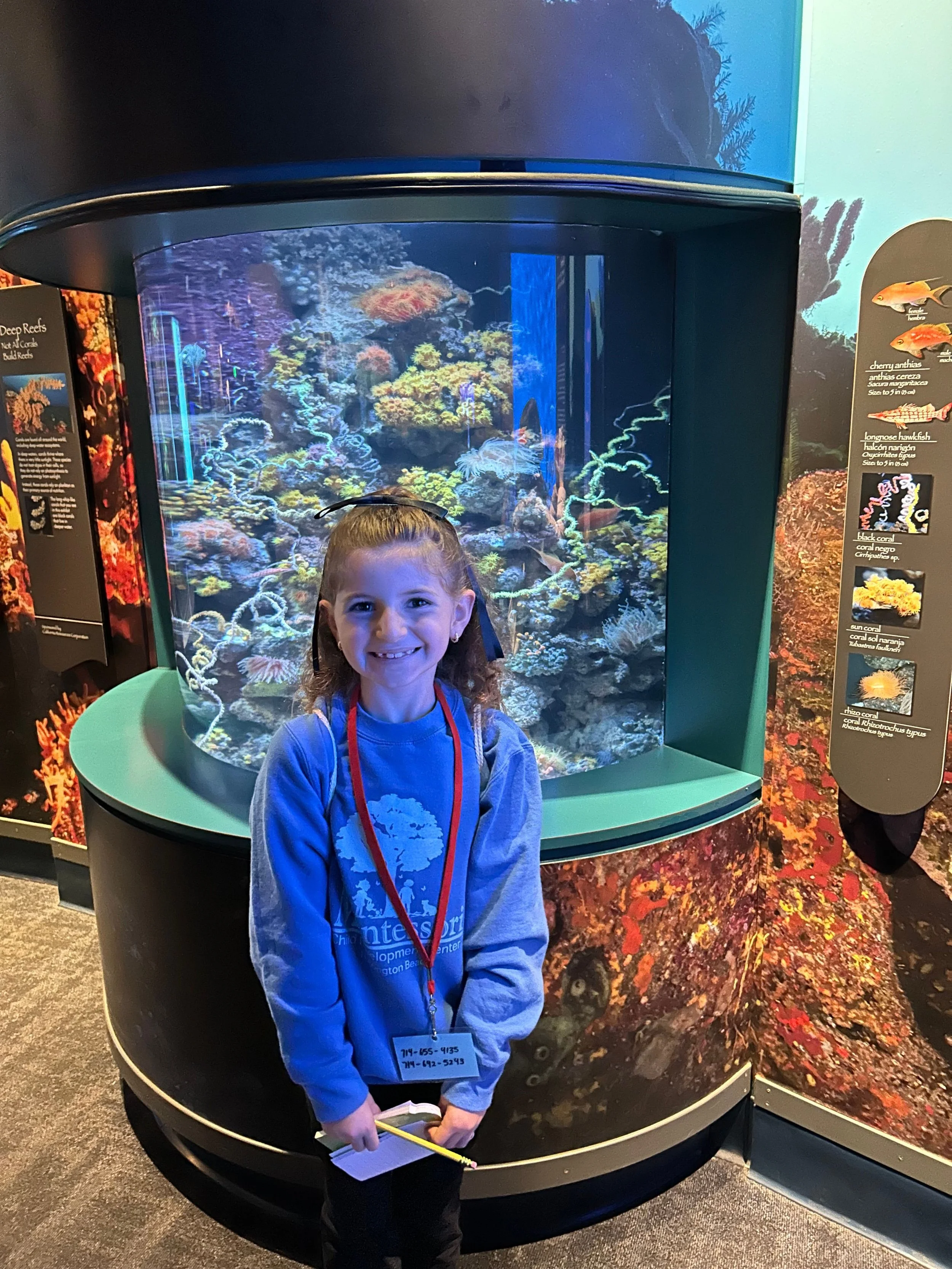 A young girl with a big smile standing in front of an aquarium display filled with colorful coral and fish.