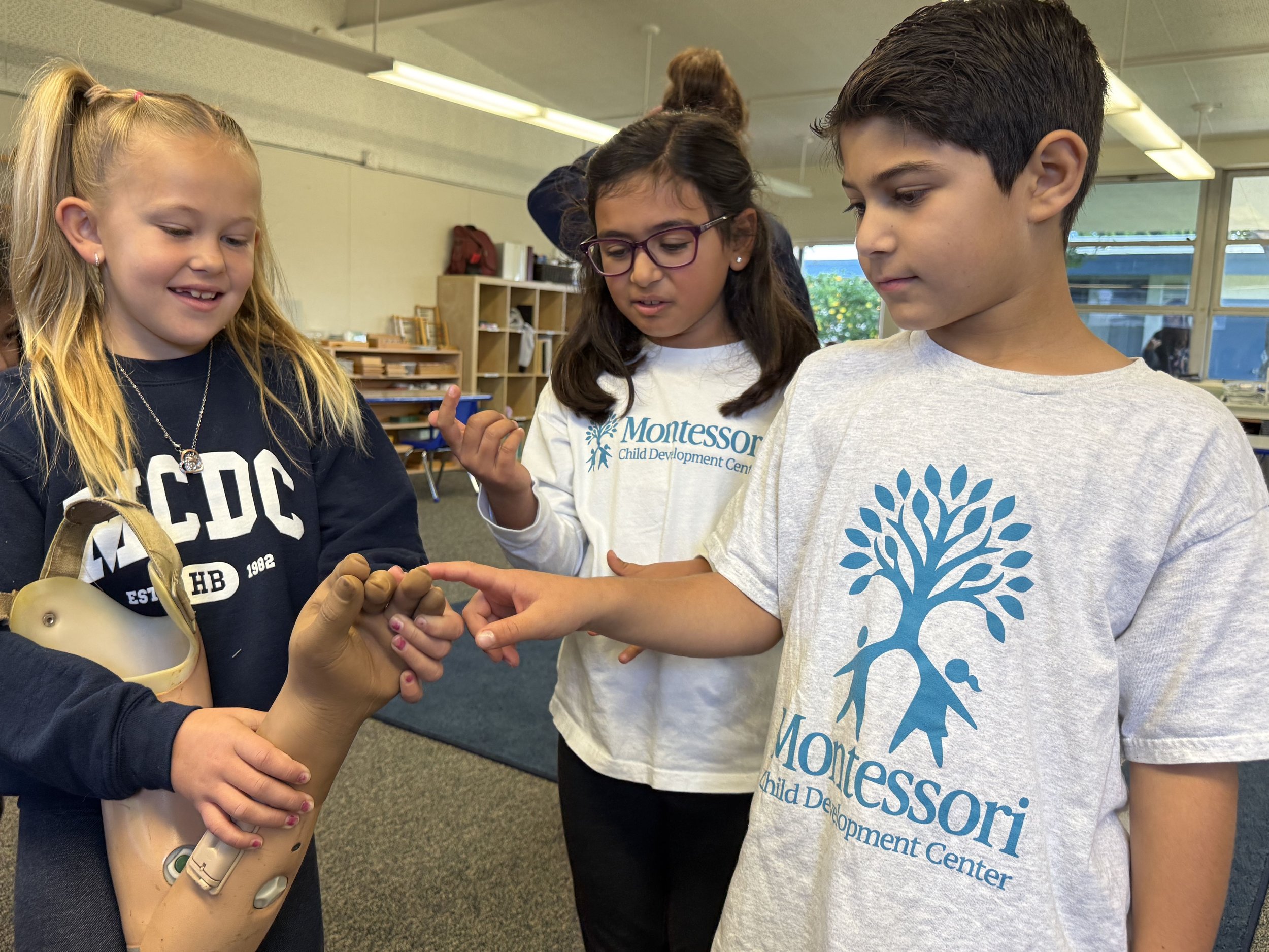 Three children, two girls and one boy, are gathered around and touching a prosthetic hand in a classroom setting.
