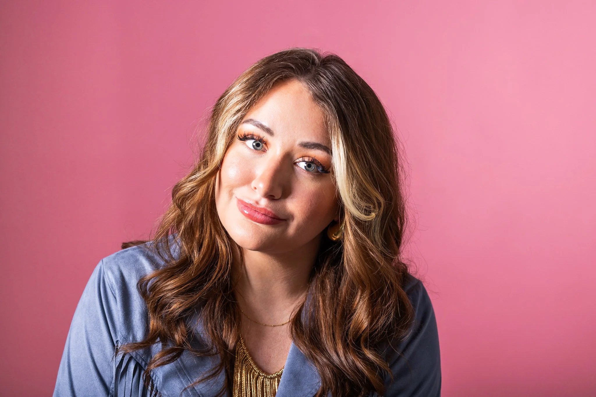 A woman with long, wavy brown hair and blue eyes looking at the camera, wearing a blue top and gold jewelry, against a pink background.