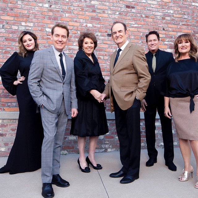 Six people standing in front of a brick wall, smiling at the camera. They are dressed in business or semi-formal attire, with some men in suits and women in dresses.