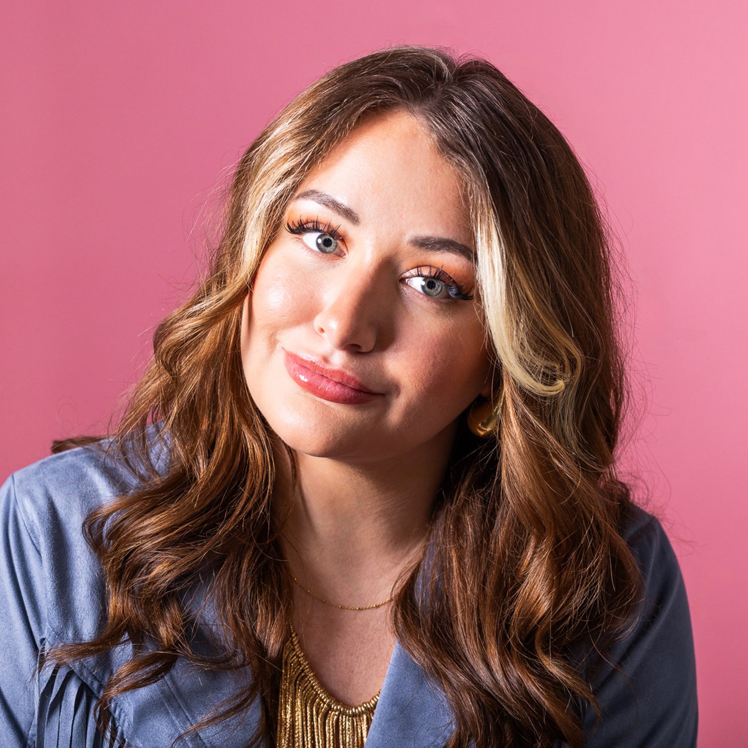 A woman with wavy brown hair, blue eyes, and makeup, wearing a blue top and gold jewelry, sits against a pink background.