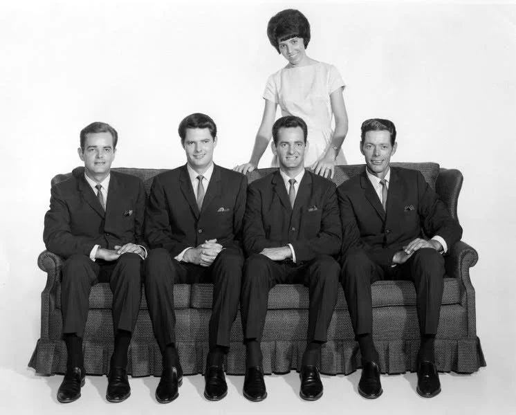 A black-and-white photo of four men in suits sitting on a couch, with a woman standing behind them with her hands on their shoulders.