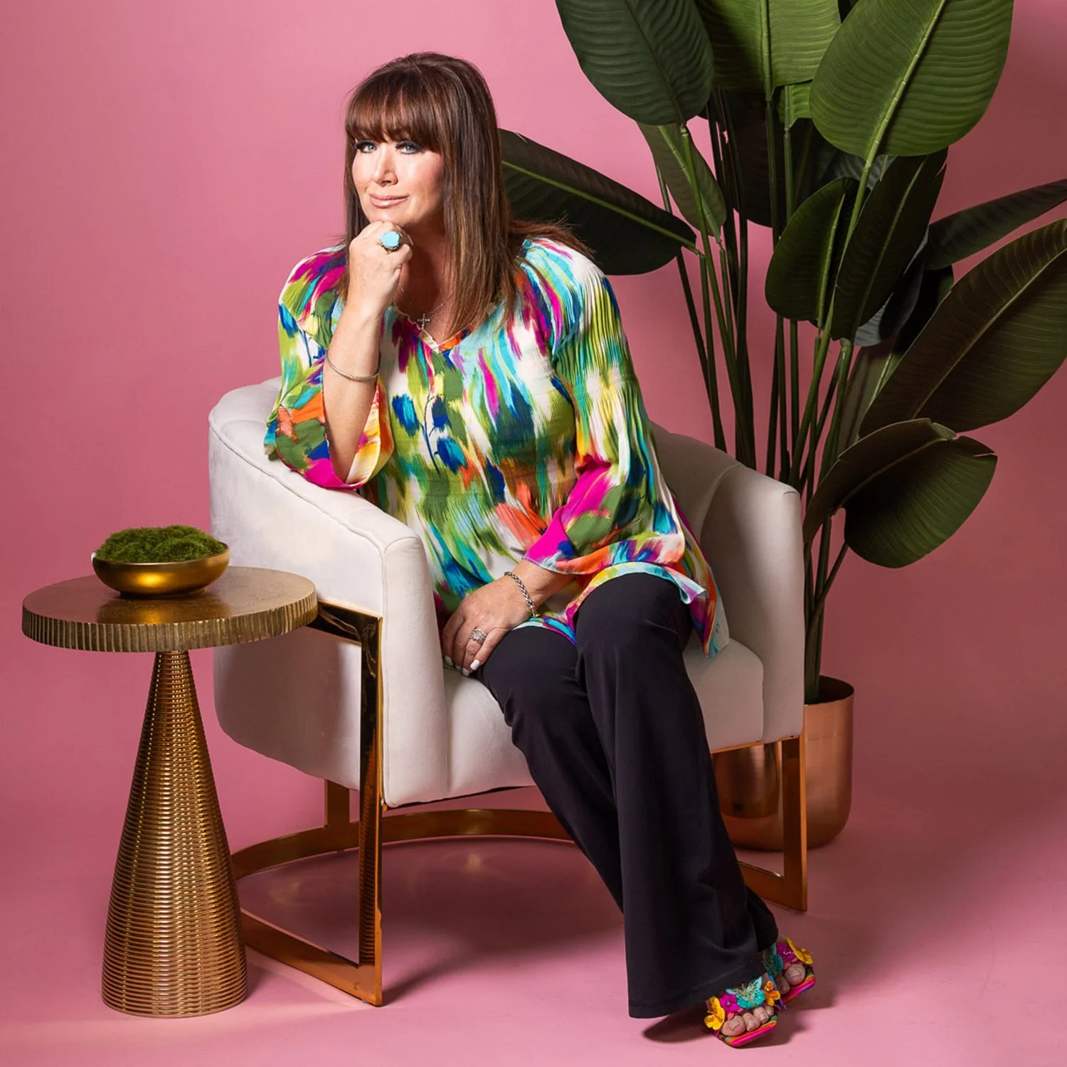 A woman with brown hair and bangs sits on a white armchair against a pink background, wearing a colorful abstract-patterned blouse and black pants. She rests her chin on her left hand, which is adorned with rings and a bracelet, and looks at the camera. There is a gold and wood side table with a small bowl of moss in front of her, and a large green potted plant behind her to the right.