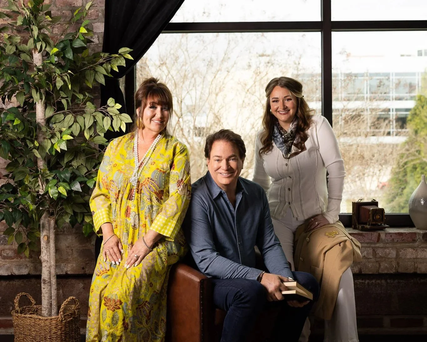 Three people smiling indoors in front of a large window, with a woman in a yellow patterned dress on the left, a man in a blue shirt sitting on a brown chair in the middle, and a woman in a white blouse on the right, with a green plant and decorative items in the background.