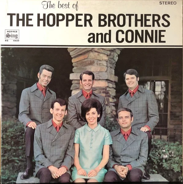 Photograph of The Hopper Brothers and Connie, a music group, posing outdoors in front of a stone archway, with six members, five men in matching gray suits and red shirts, and one woman in a light blue dress.