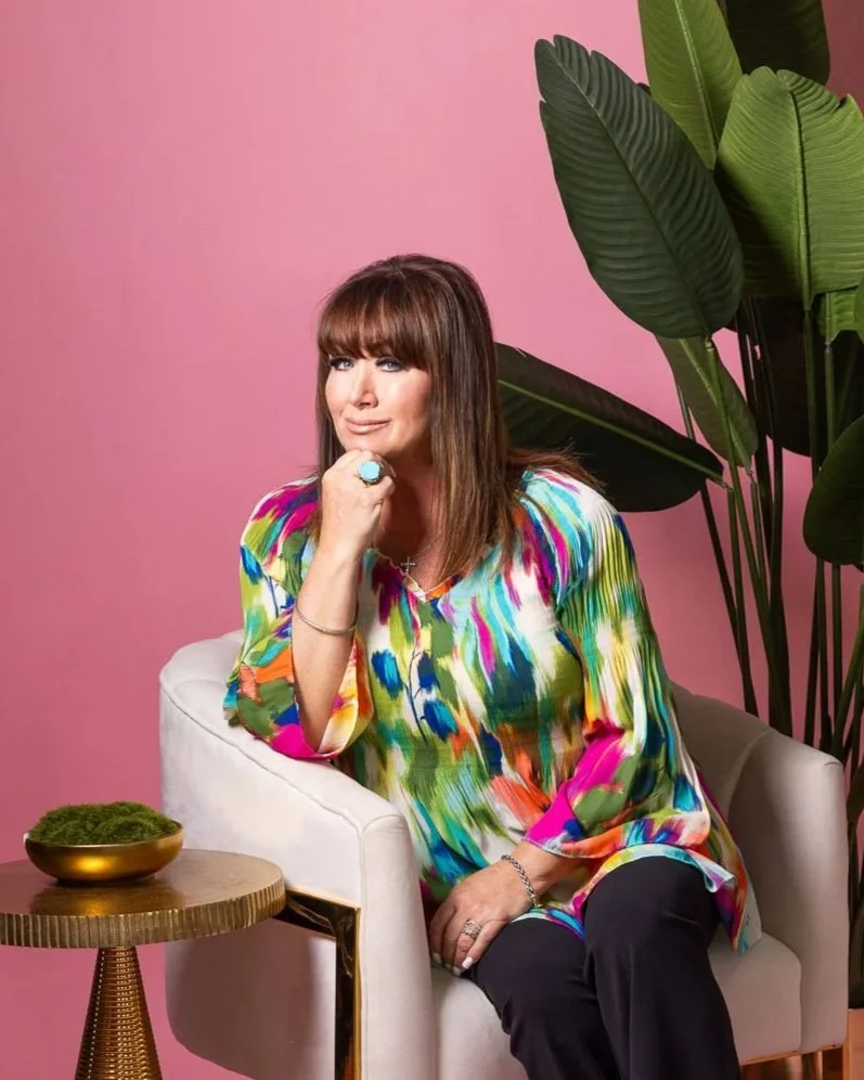 A woman with brown hair and bangs sitting on a white armchair against a pink background, wearing a multicolored abstract top. She has her right hand resting on her chin, showing a ring with a large turquoise stone, and is smiling slightly. There is a small round table with a golden base and a green plant in a gold bowl next to her.