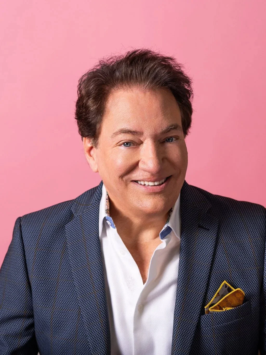 A smiling man with dark brown hair wearing a dark blue patterned blazer and white shirt with a yellow pocket square, against a pink background.