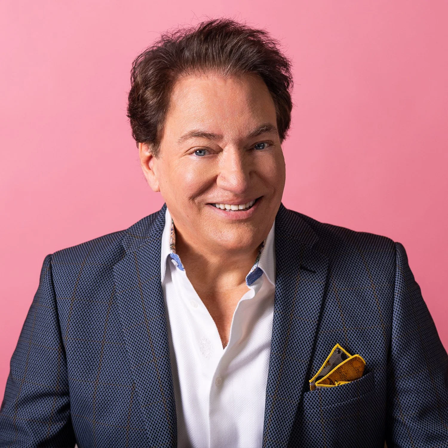 A smiling man with dark hair and blue eyes wearing a navy checkered blazer and white shirt, with a yellow pocket square, against a pink background.