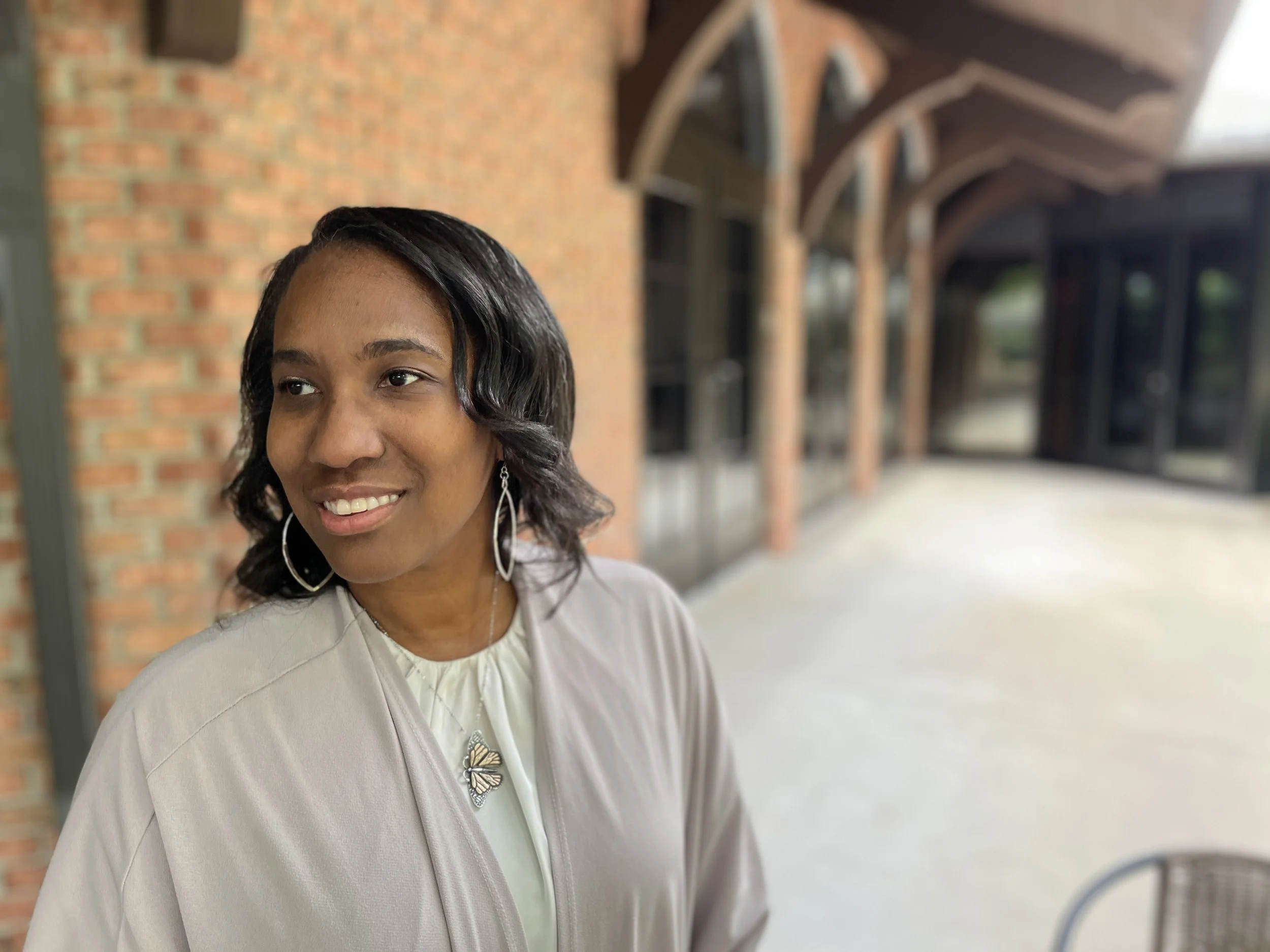 A woman with shoulder-length black wavy hair smiling, wearing earrings, a butterfly-shaped brooch, and a light-colored jacket, standing outdoors in front of a brick building with a covered walkway.