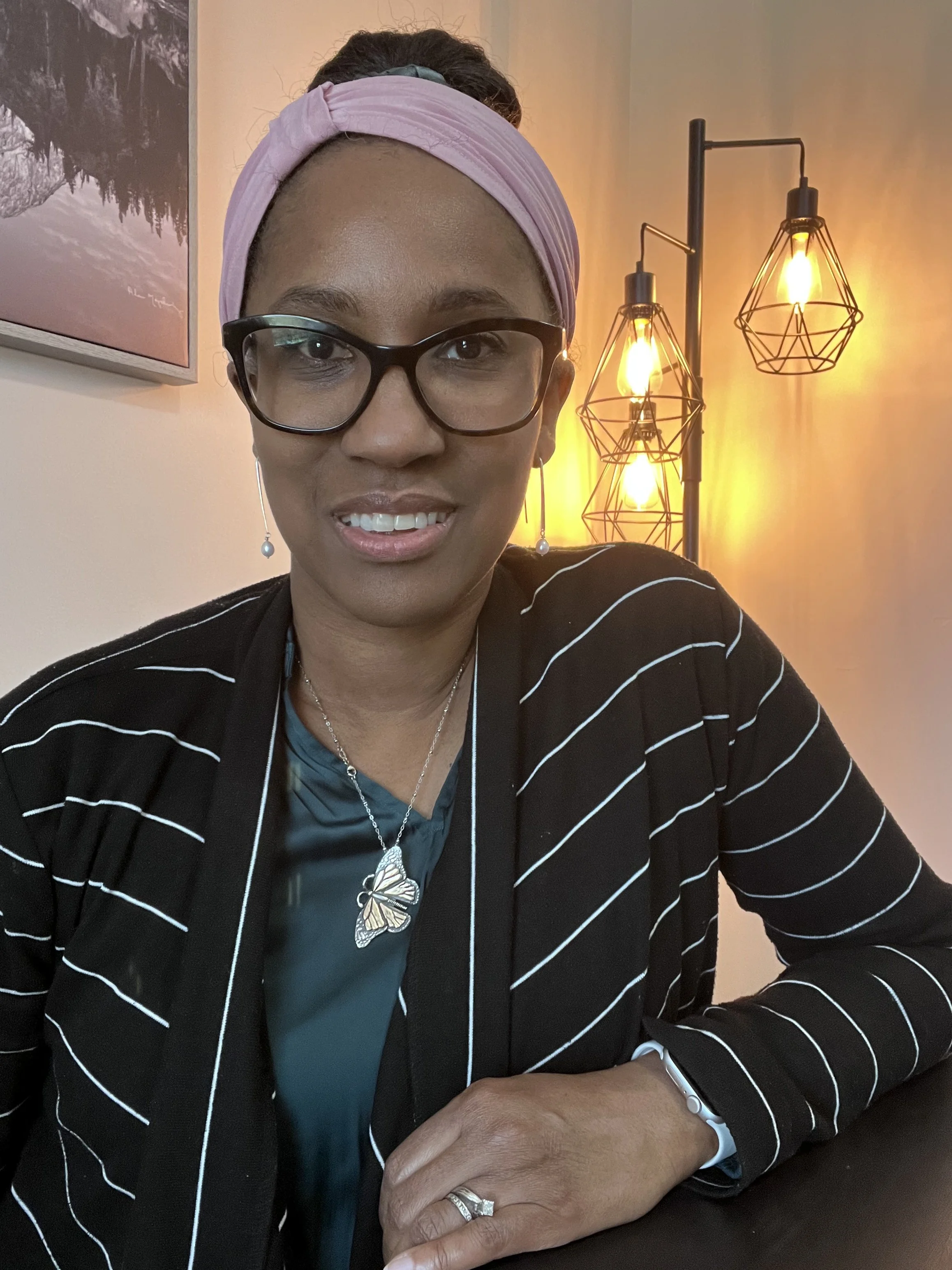 A woman wearing glasses, a pink headband, a butterfly pendant necklace, and a ring, smiling at the camera, in a room with warm lighting and modern decor.