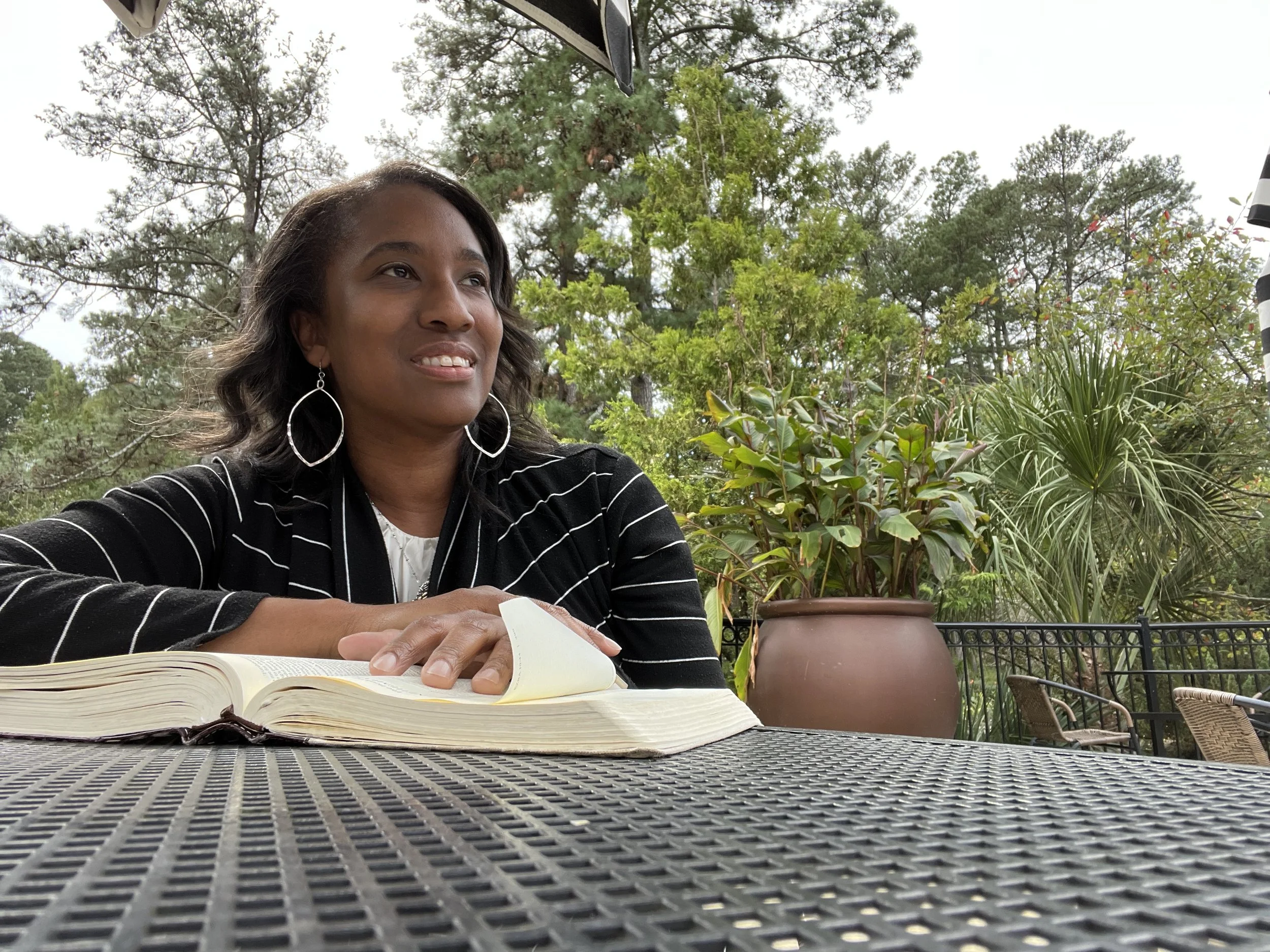 A woman sitting outdoors at a table with a book open in front of her, surrounded by lush green plants and trees.