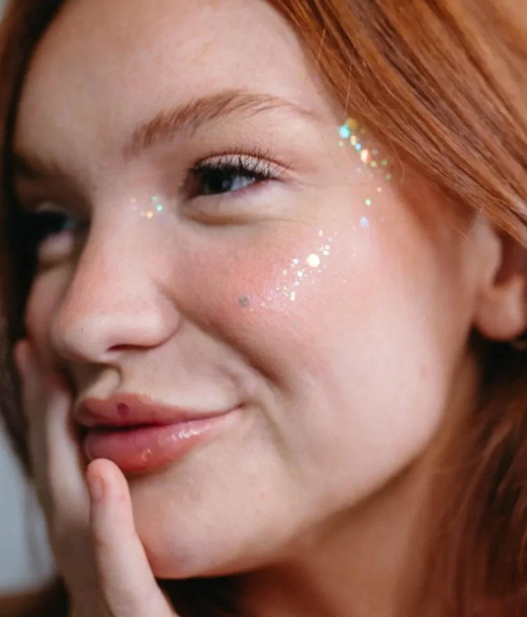 Close-up of a woman with red hair and glitter on her cheek, touching her lips with her finger, smiling slightly.