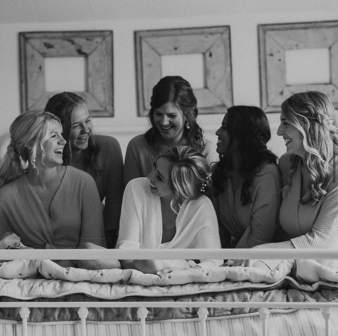 Black and white photo of seven women laughing and smiling while sitting on a bed, dressed in robes, in a room with simple wall art.
