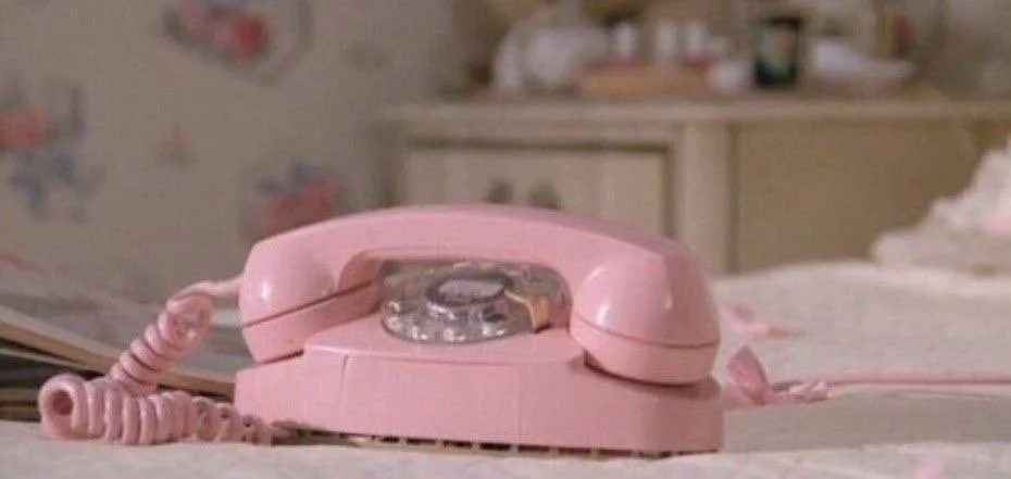 A pink rotary dial telephone on a bed in a room with pastel-colored decor.
