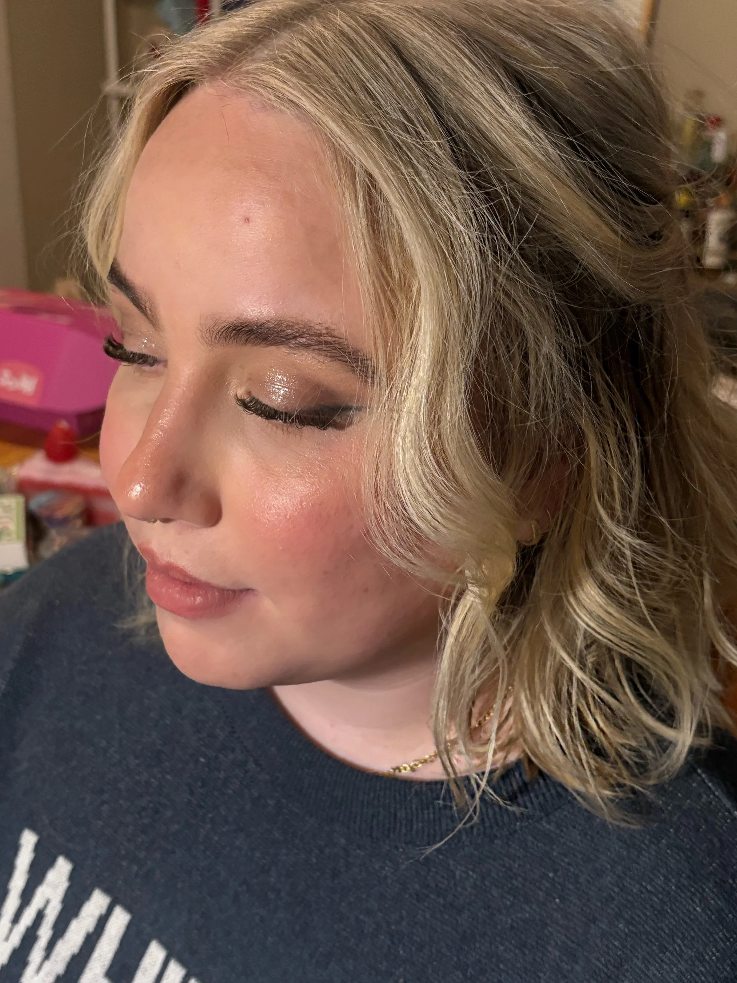 A woman with blonde, curly hair and closed eyes wearing makeup, including glittery eyeshadow and glossy lips, dressed in a dark top.