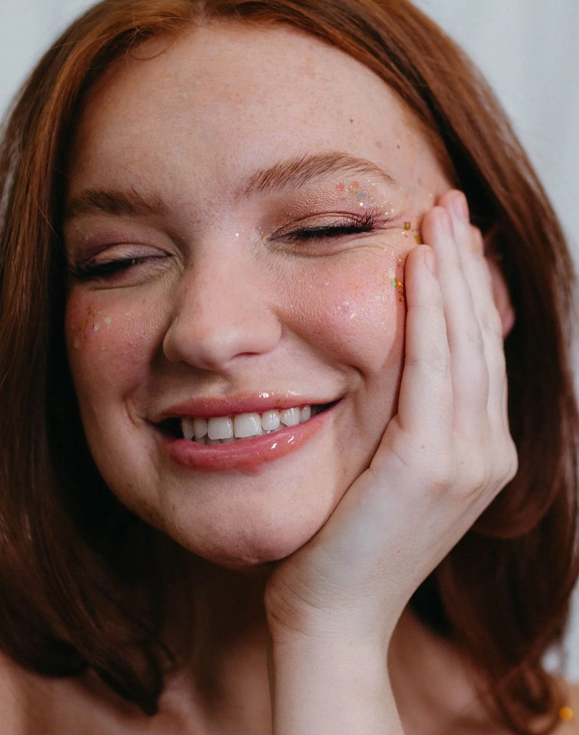 Close-up of a smiling woman with red hair, her eyes closed, wearing pink glitter makeup on her eyelids and cheeks, and with her hand gently touching her face.