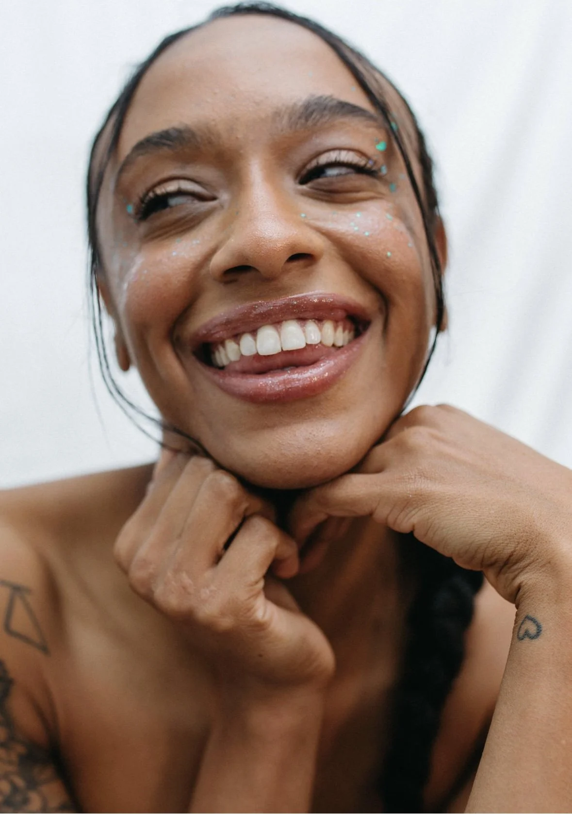 Close-up of a smiling woman with glitter makeup, tattoos, and wet hair, resting her chin on her hands.