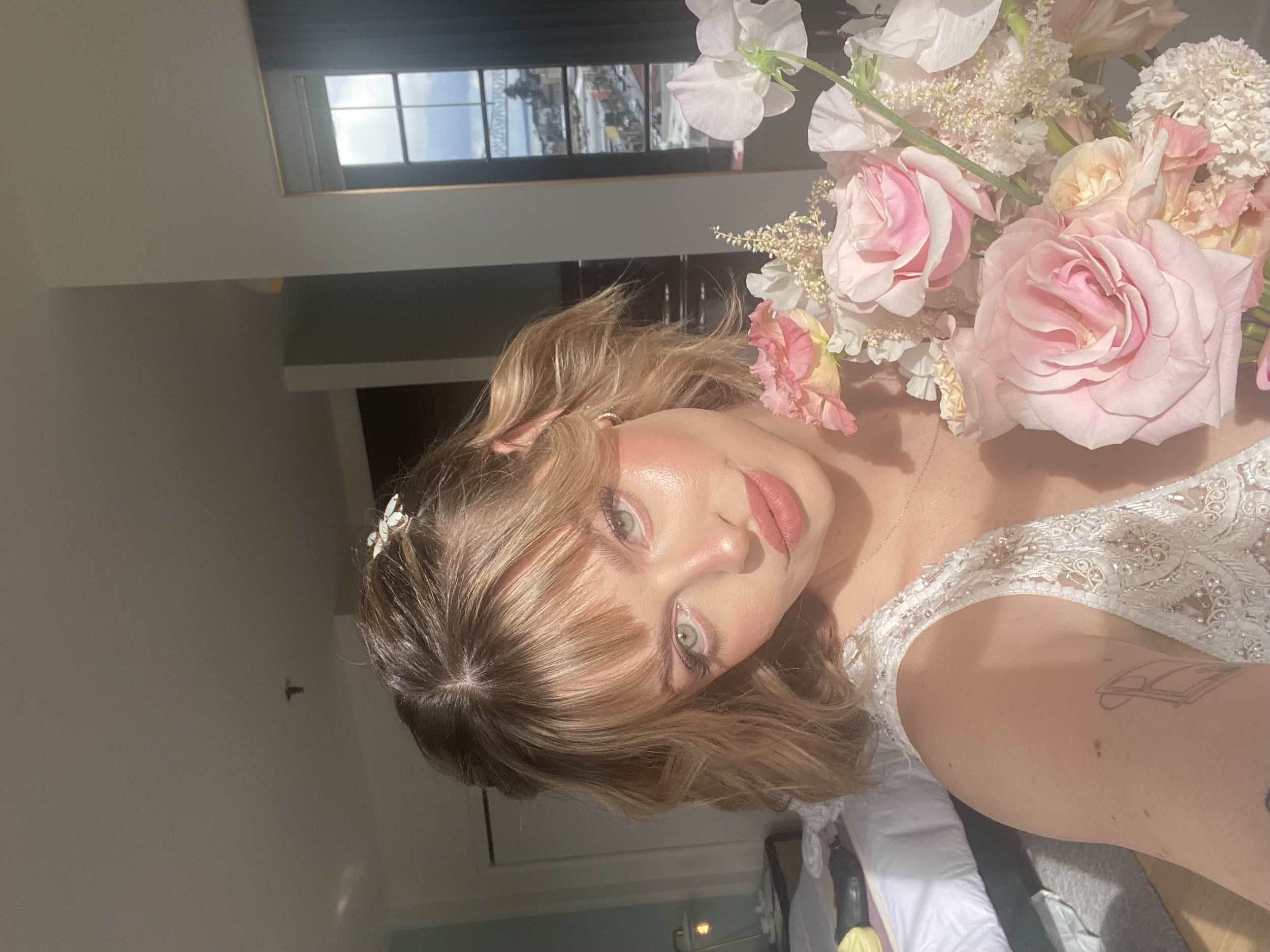 A woman with wavy blonde hair and light makeup taking a selfie in a room with a large bouquet of pink and white flowers, wearing a white lace dress with short sleeves.