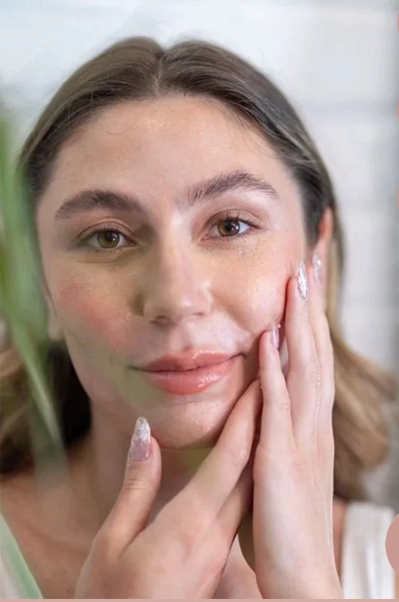 A woman with light brown hair applying a skincare product or moisturizer to her face with her fingers.