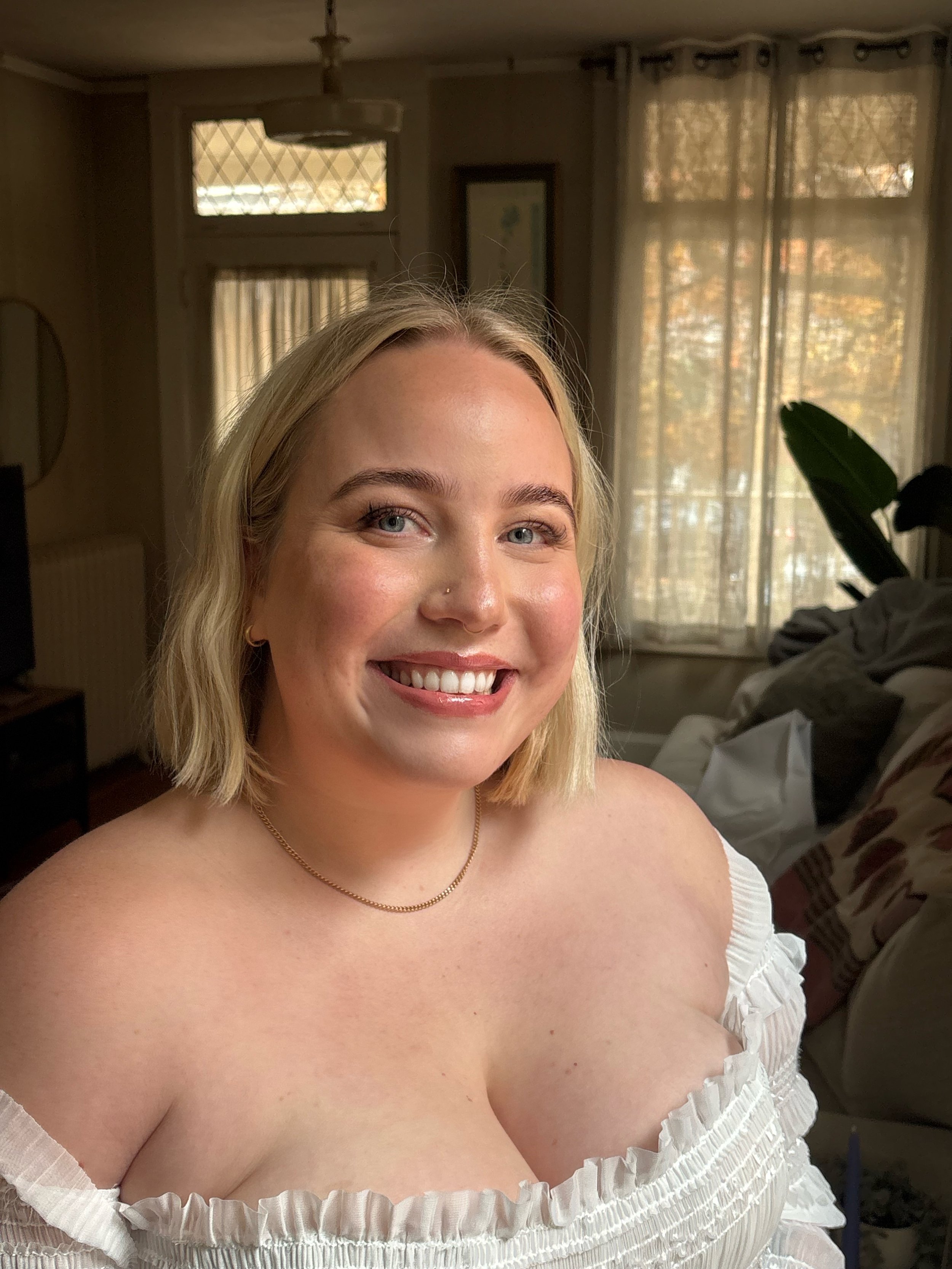 A smiling young woman with blonde hair, wearing a white off-shoulder top, sitting in a well-lit room with curtains and a plant in the background.