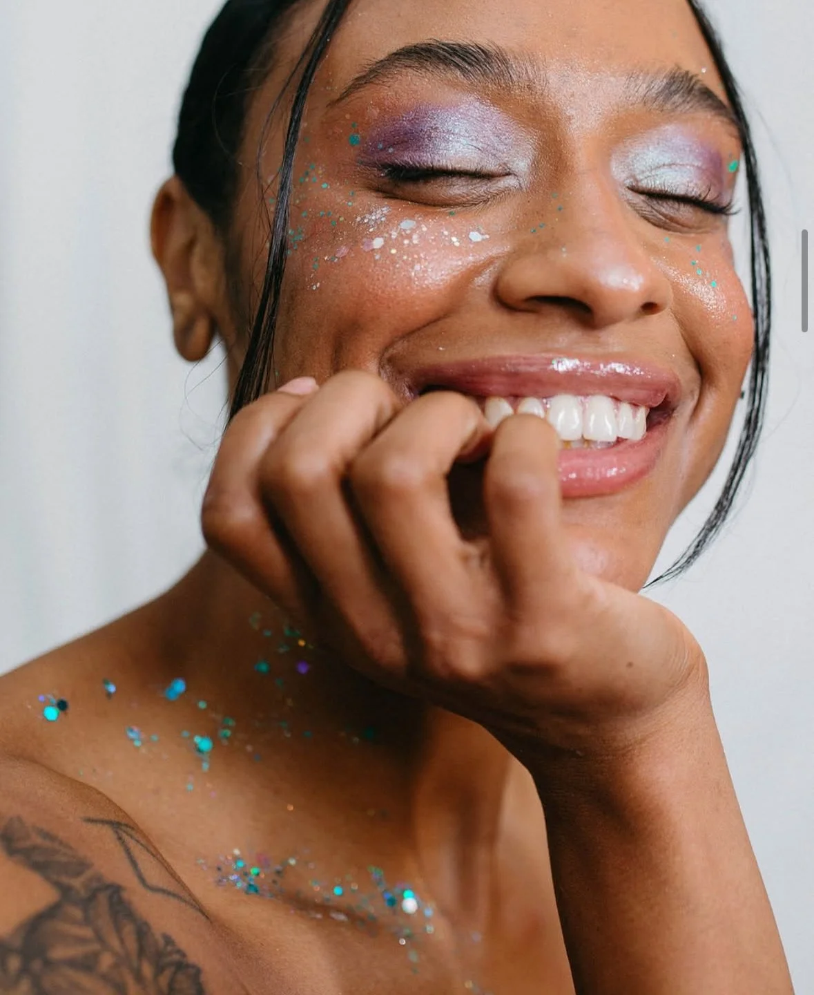 A smiling woman with glitter on her face, shoulder, and neck, wearing makeup with purple and silver eyeshadow, with one eye closed, resting her chin on her hand.