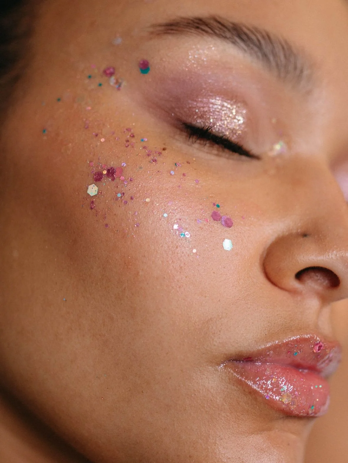 Close-up of a woman's face with glittery makeup, pink and glitter eyeshadow, and colorful glitter on her cheek and lips, with her eyes closed.