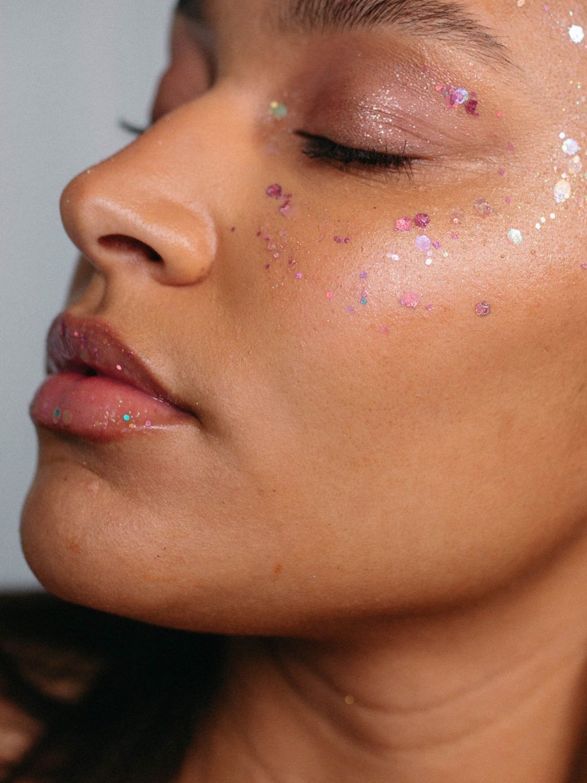 Close-up of a woman's face with closed eyes, decorated with pink, purple, and iridescent glitter on her eyelids, cheeks, and lips.