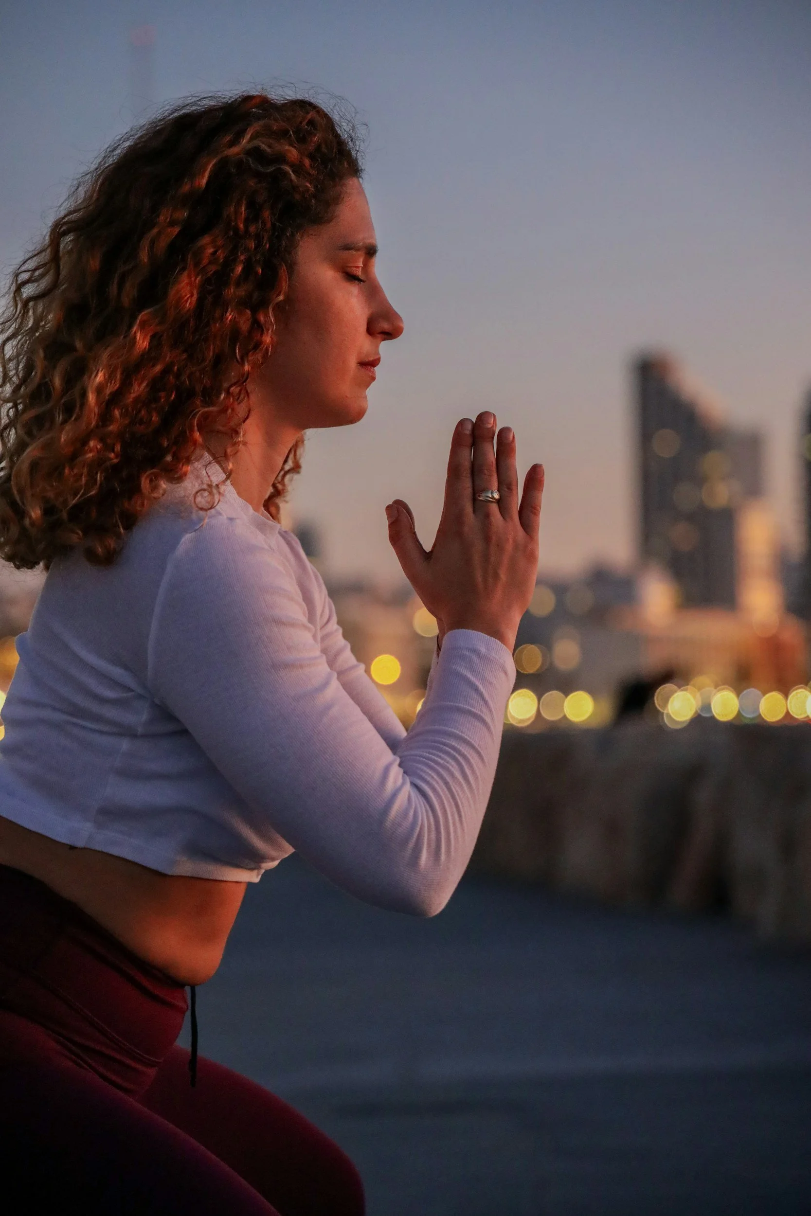 A woman with curly hair practicing yoga or meditation outdoors at sunset, with a city skyline in the background.