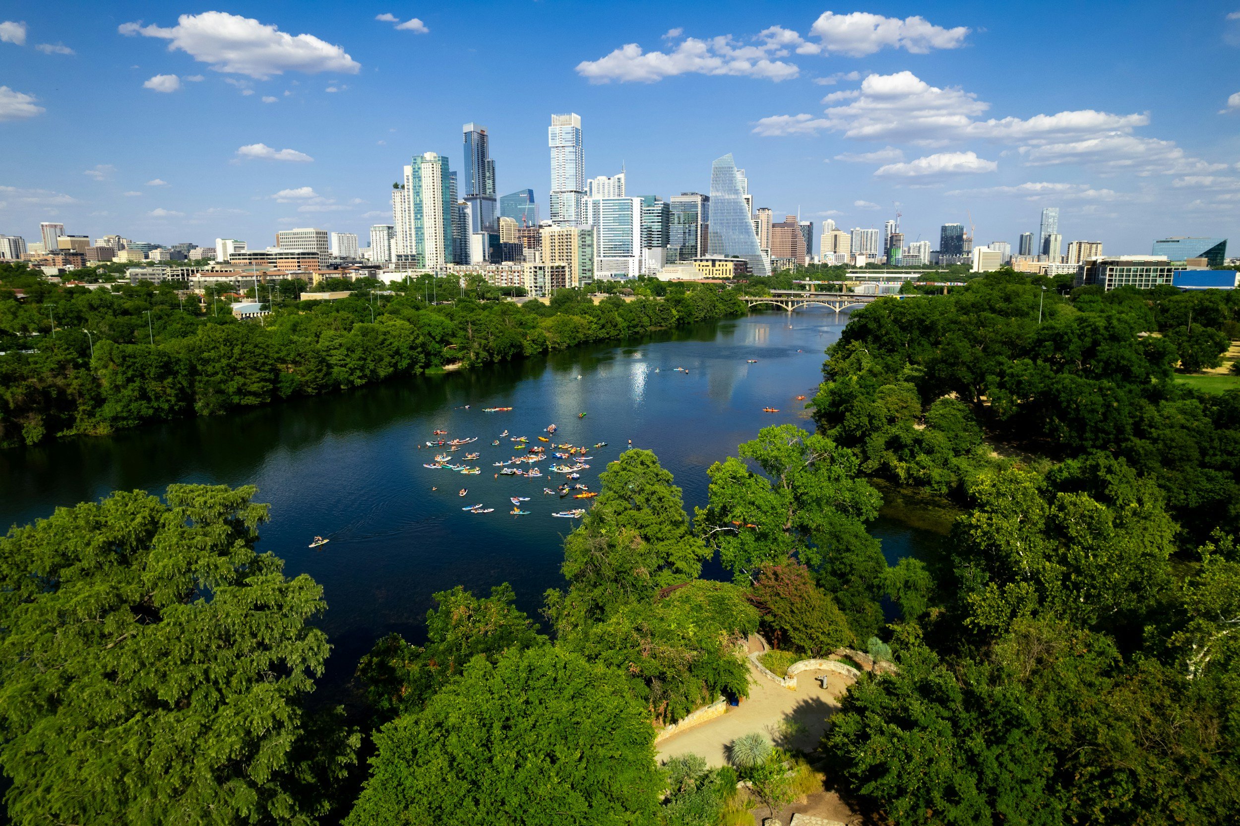 A city skyline over a river with numerous kayakers and lush green trees along the riverbank.